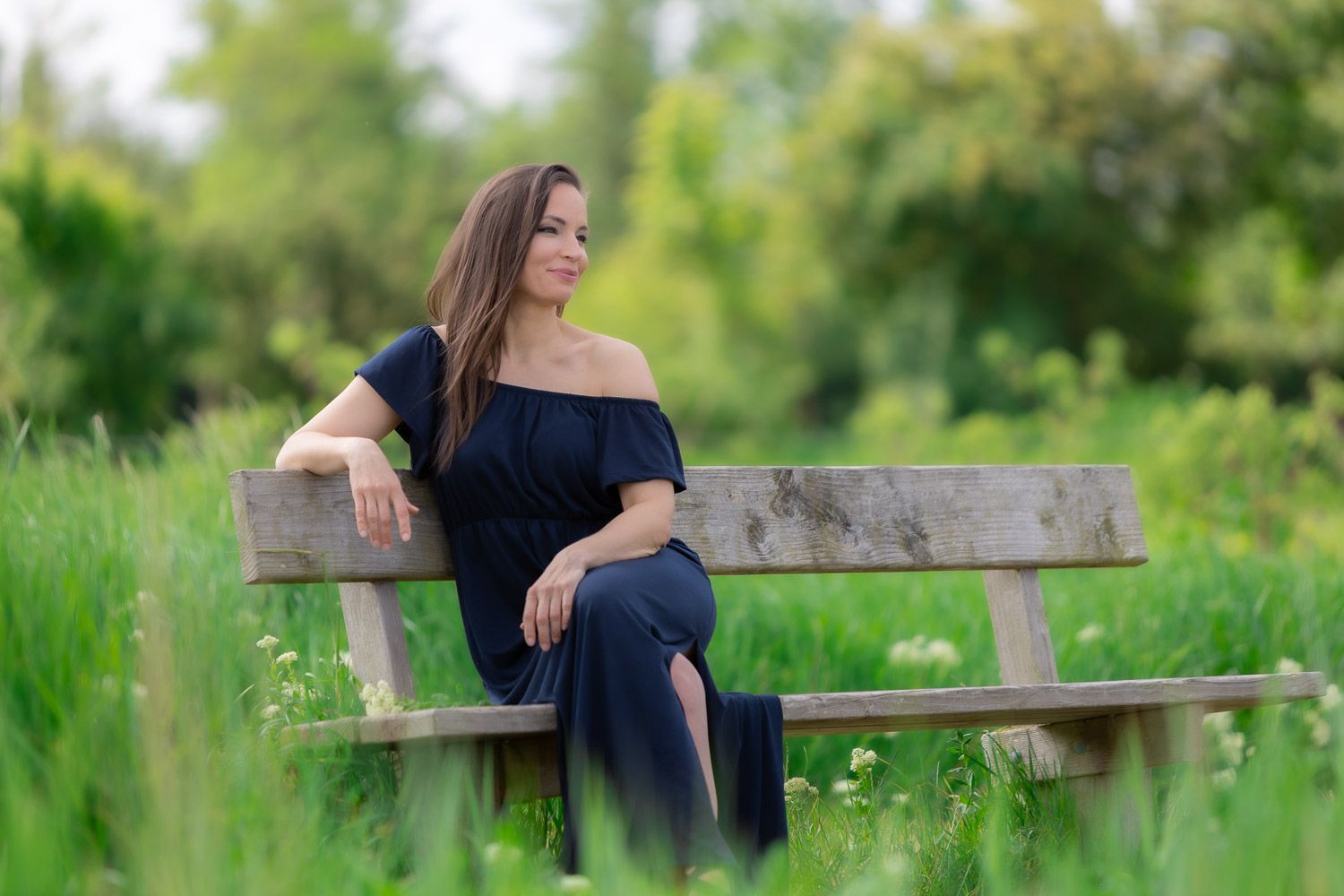 Woman in navy blue dress sitting on wooden bench surrounded by lush green grass and wildflowers.
