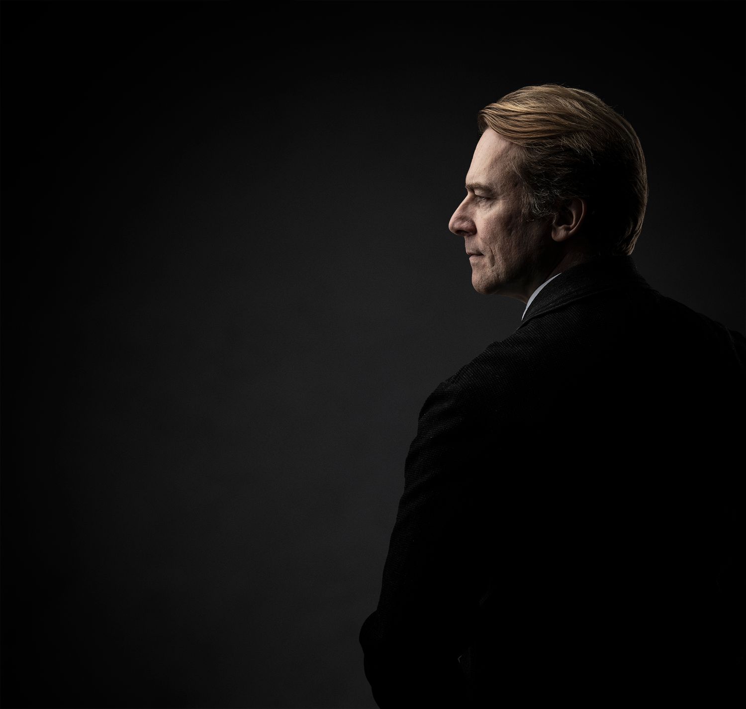 Professional portrait of person in dark suit against black background with dramatic side lighting.