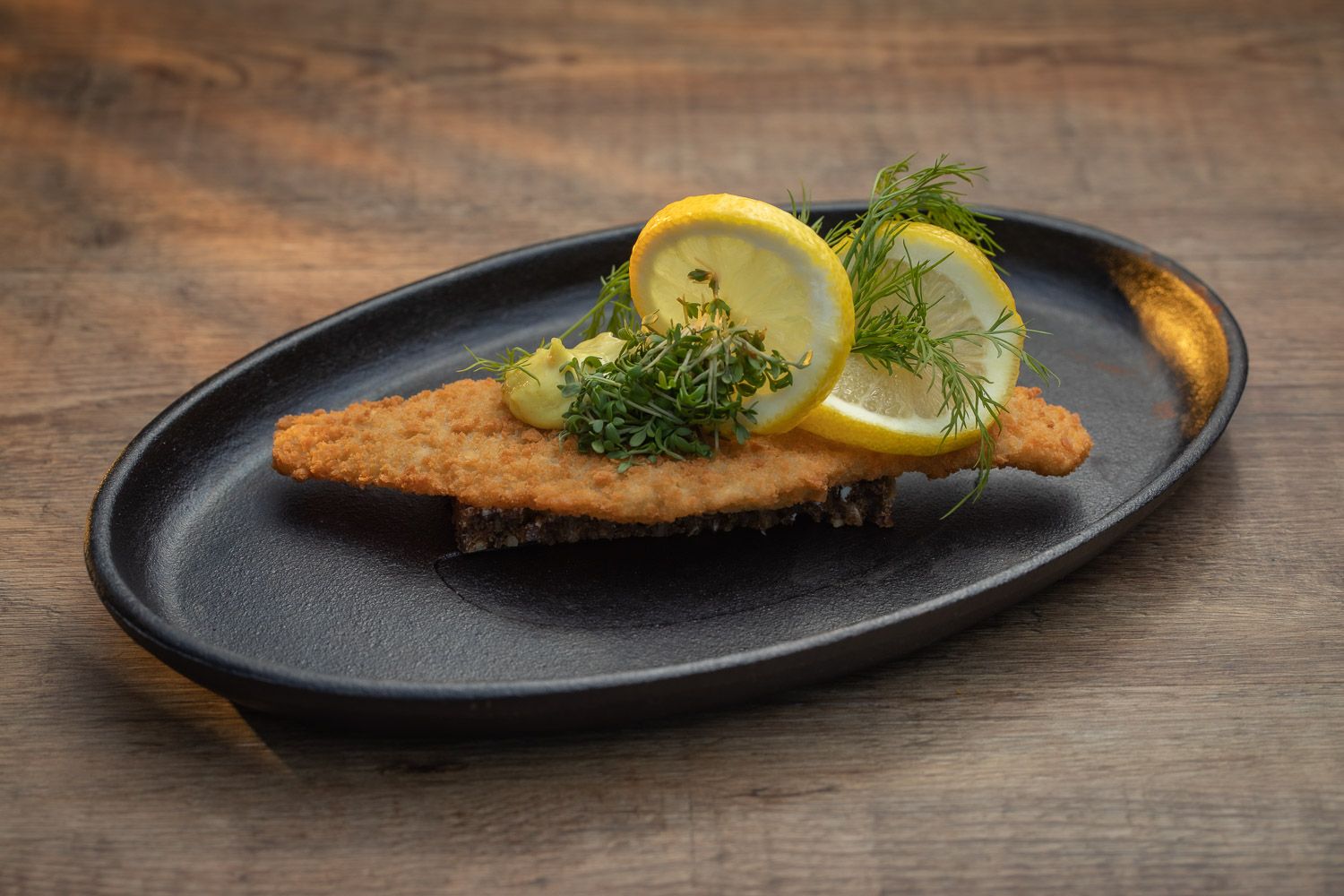 Breaded fish fillet garnished with fresh dill and lemon slices on a black plate.