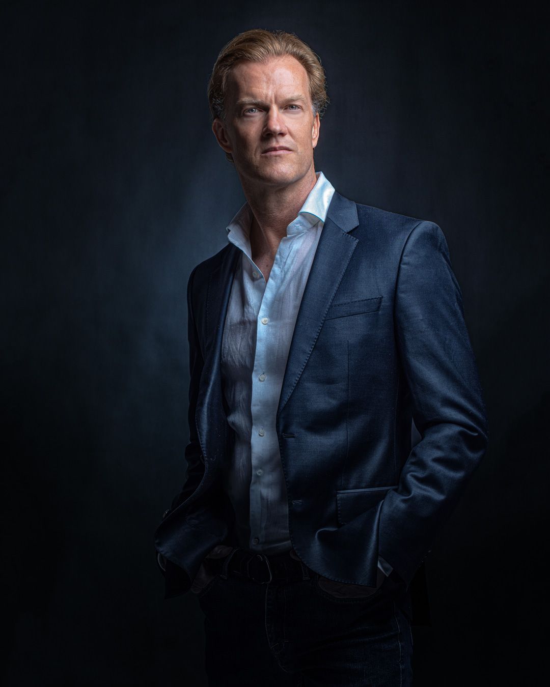 Professional headshot of person in navy suit with open collar against dark background.