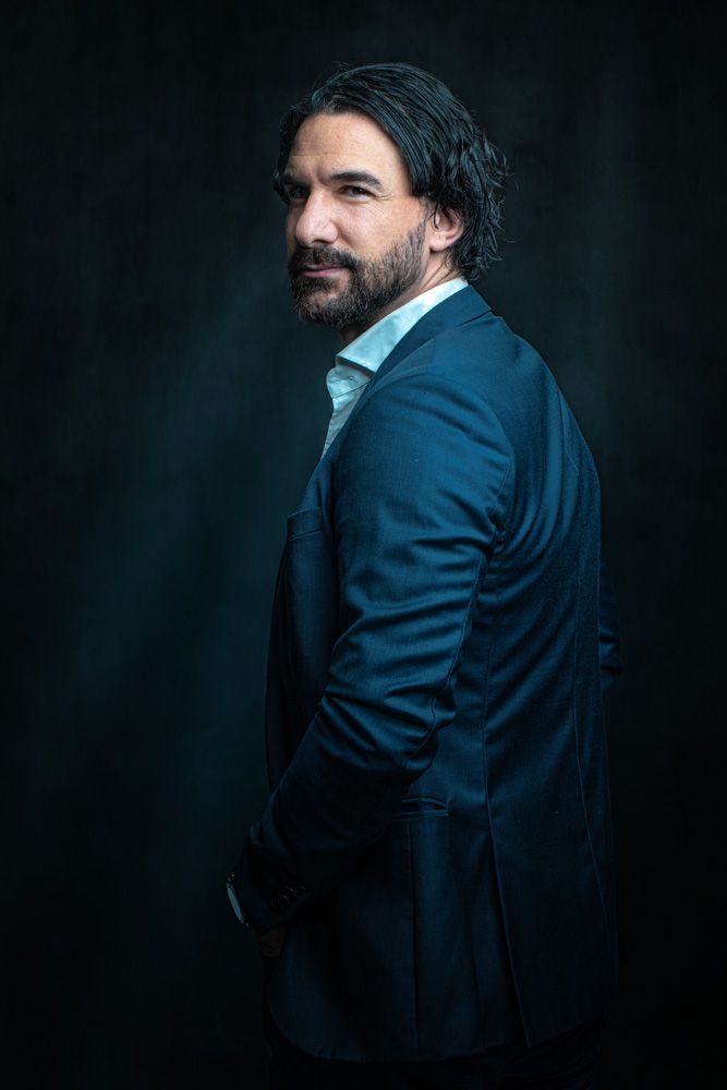 Dramatic studio portraits in blue suit against dark background with moody lighting.