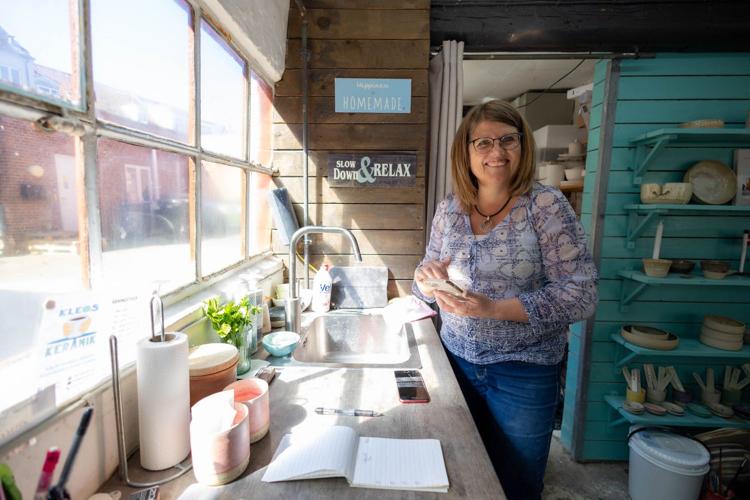 A pottery studio workspace with shelving and supplies near a bright window.