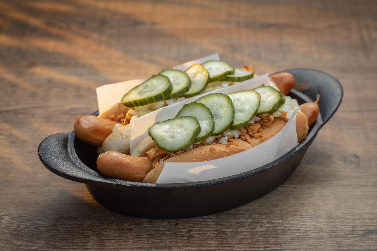 Hot dog with fresh cucumber slices and toppings served on dark plate.