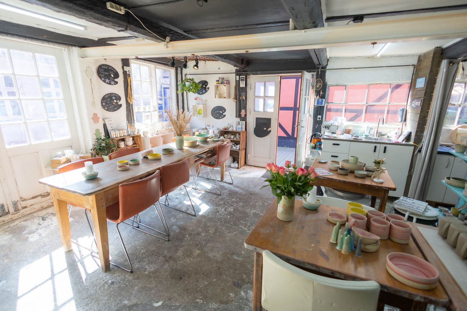 Sunlit pottery studio with wooden tables and hanging plants displays various ceramic tools and equipment.