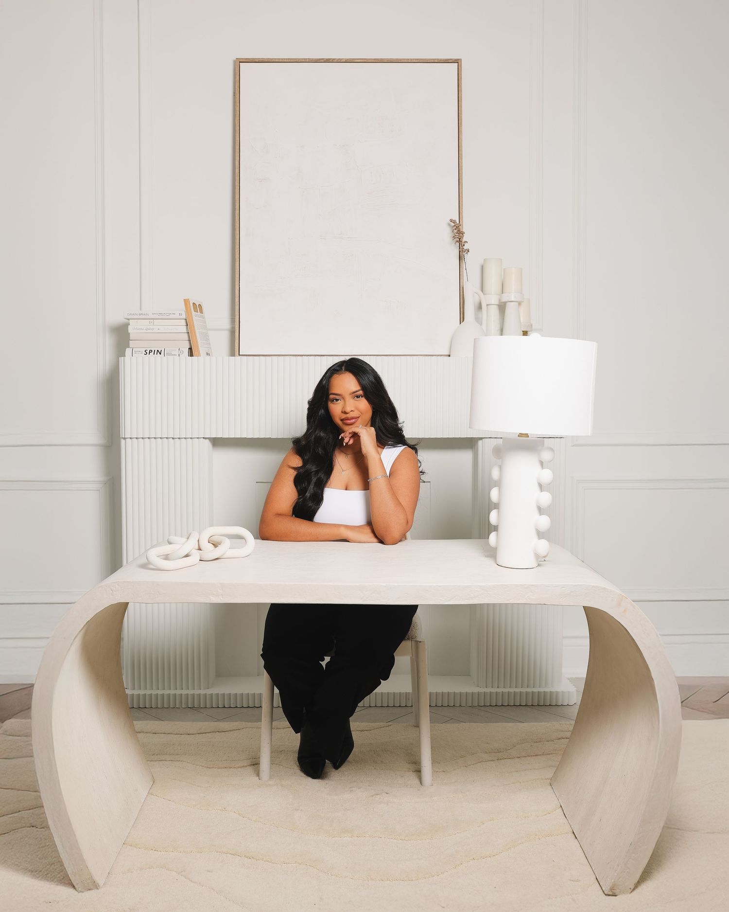 Woman sitting at a modern white curved desk in an elegant, minimalist white room with a lamp and framed art.