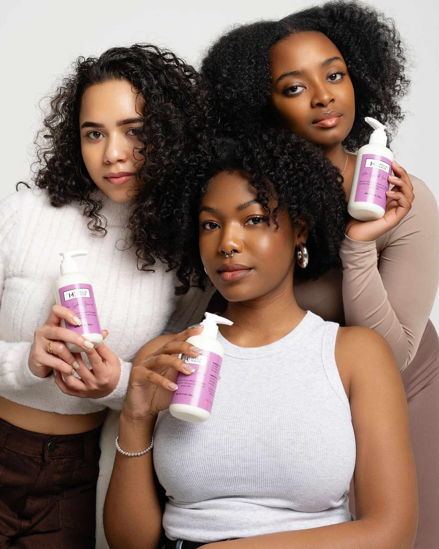 Three women with natural curly hair pose together holding purple hair care product bottles against a white background.