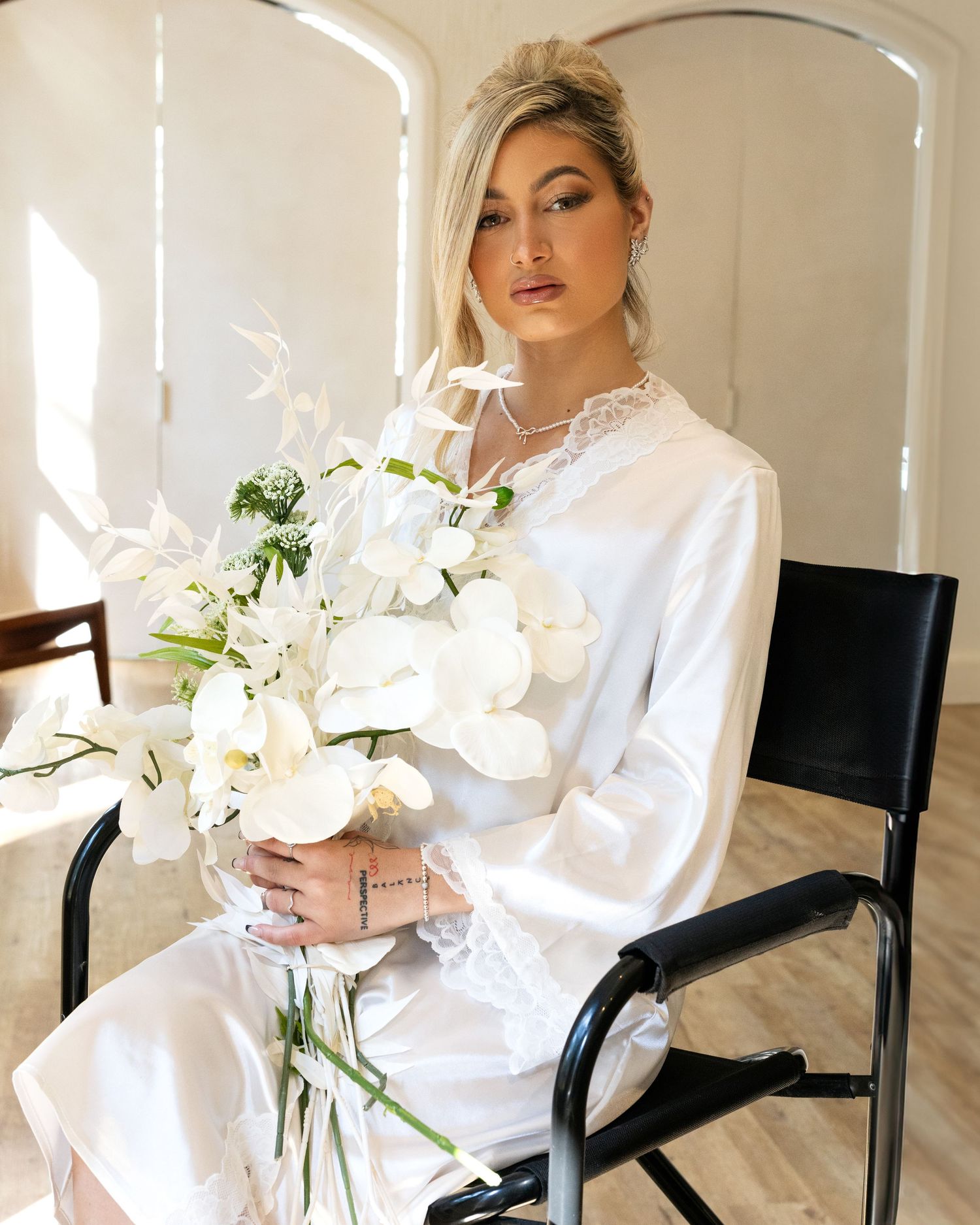 Blonde woman in white satin robe sits on black chair holding elegant white orchid bouquet in bright minimalist room.