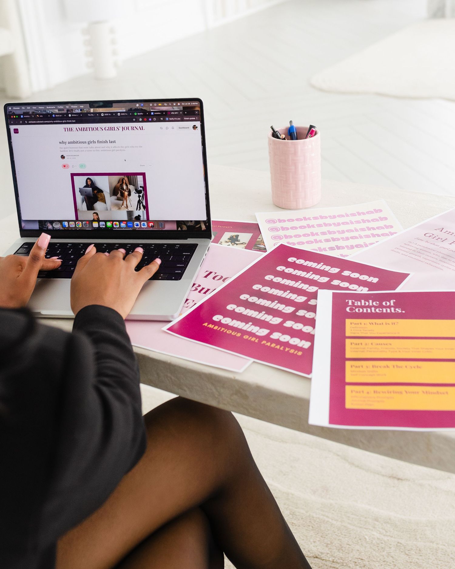 Woman working on laptop at desk with pink and yellow marketing documents and a pencil cup nearby.