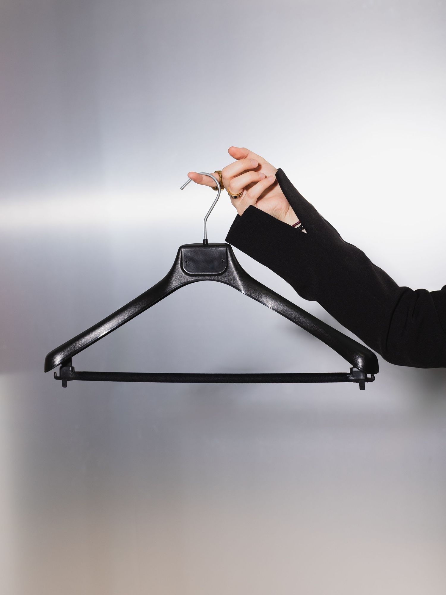 A hand in a black sleeve holds a black plastic clothes hanger against a gray background.