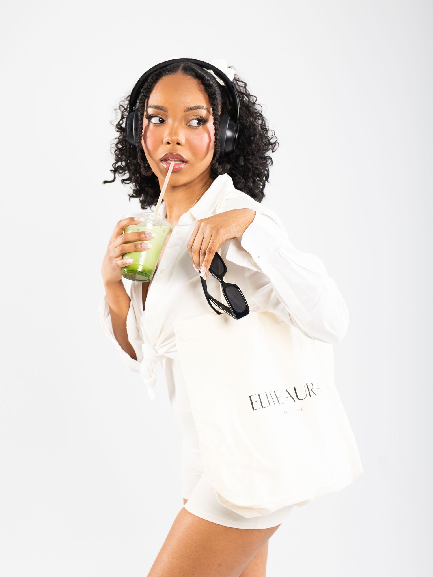 Woman in white outfit sipping green drink, wearing headphones, holding ELILENUR tote bag on white background.