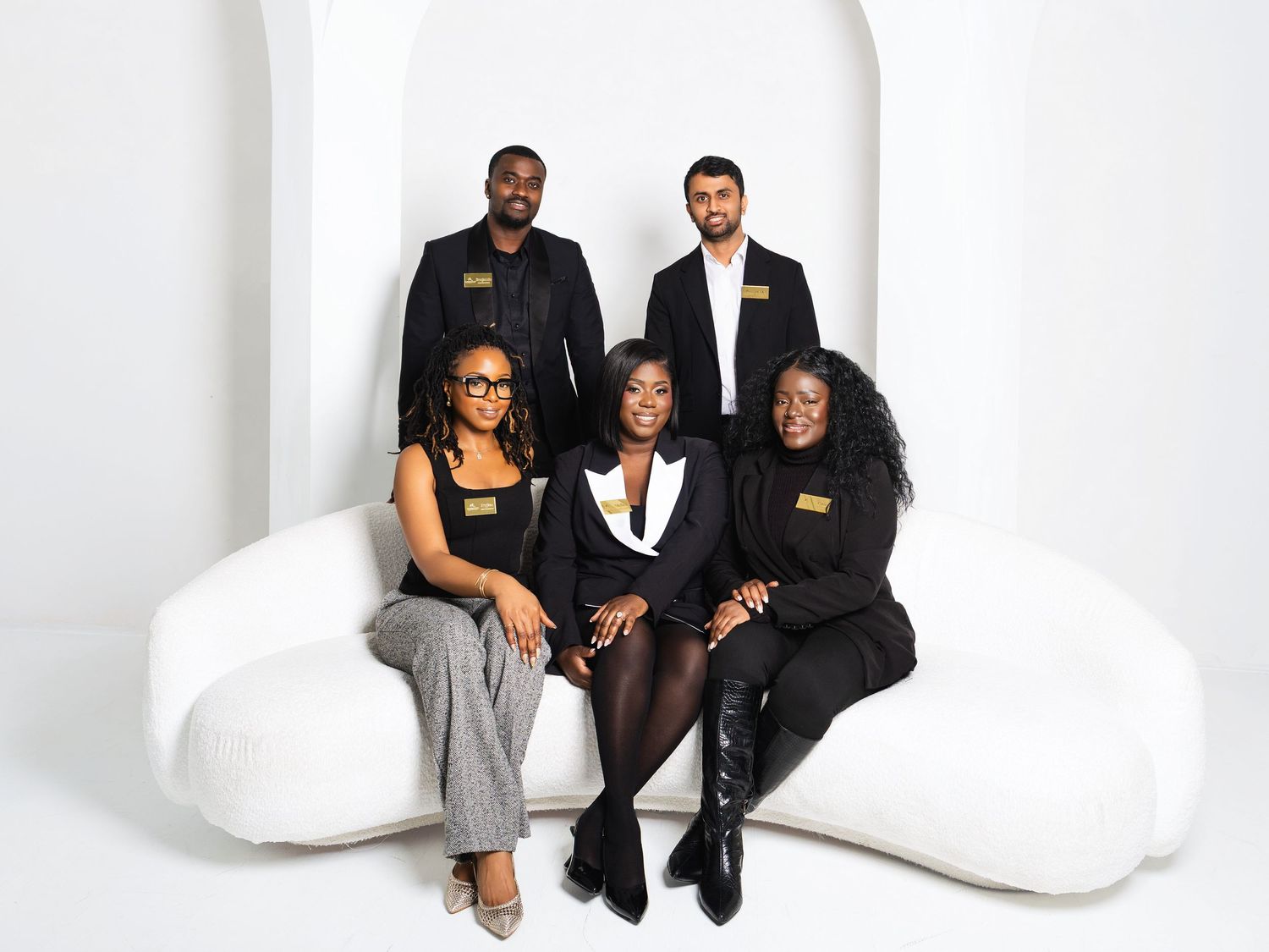 Five diverse business professionals in black outfits seated and standing on white sofa.