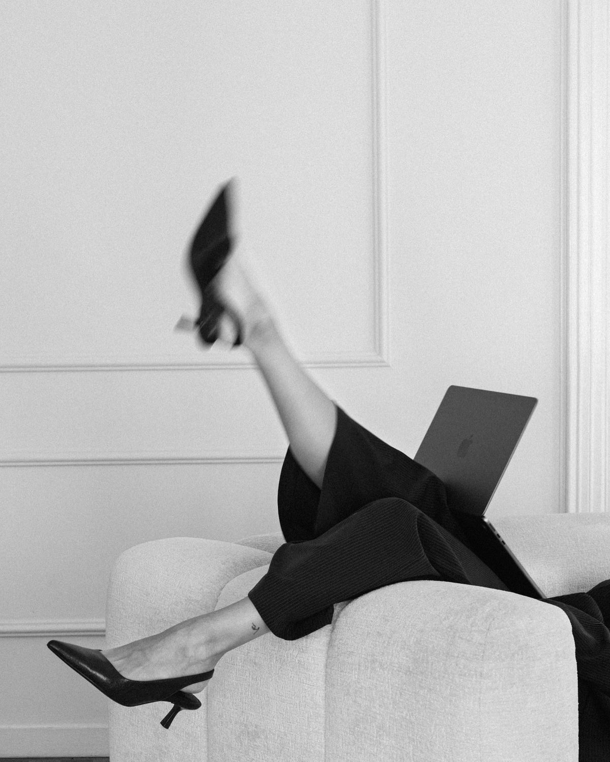 Black and white photo of a woman in heels reclining on a sofa with a laptop, legs raised elegantly.