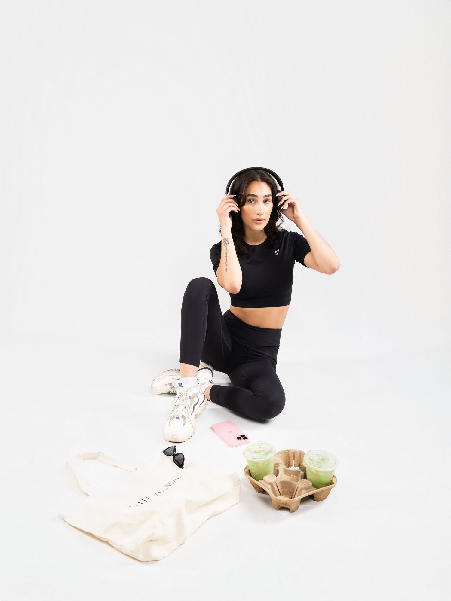 Woman in black crop top and leggings sits on floor with headphones, tote bag, and accessories nearby.