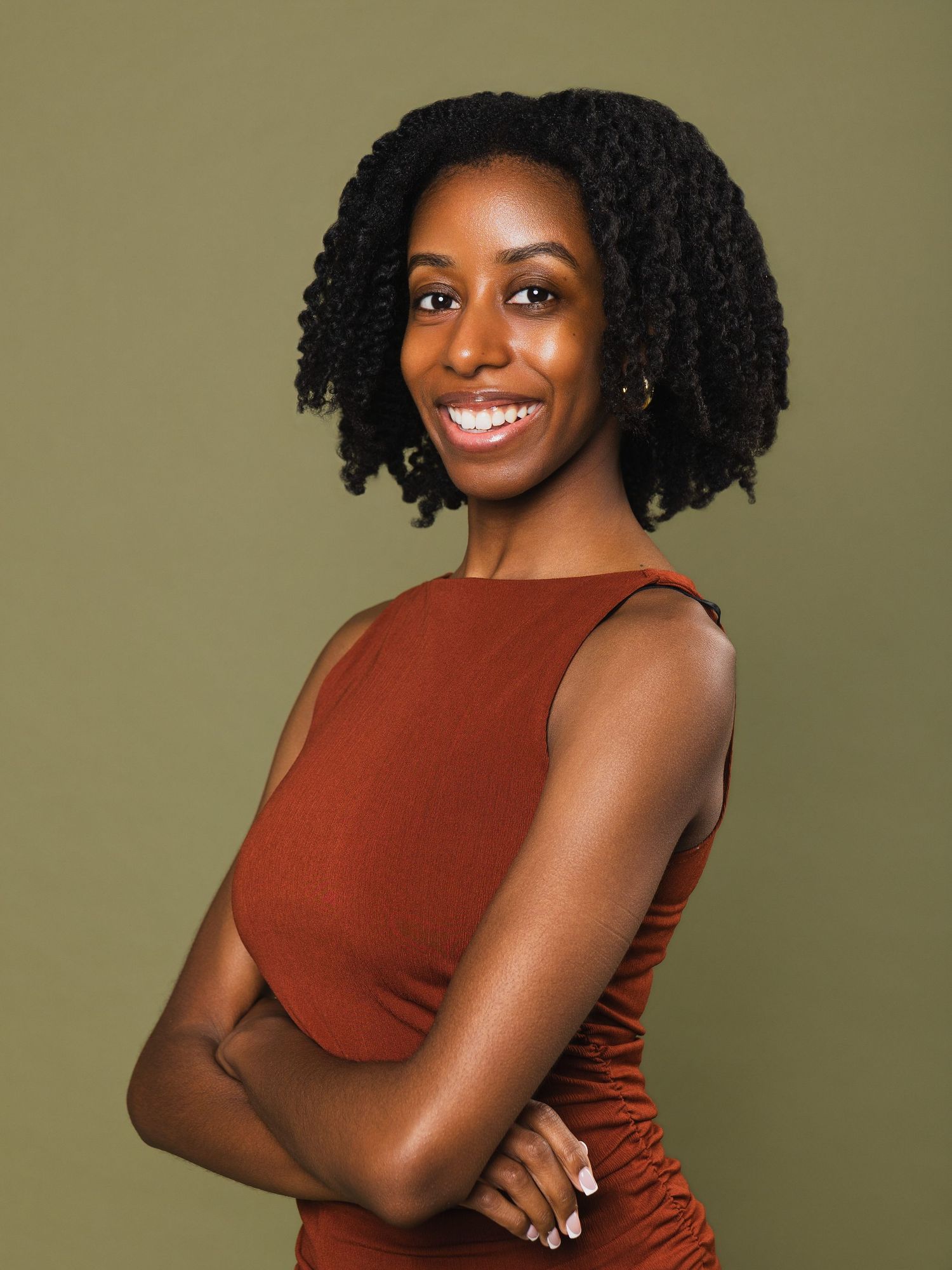 Confident young Black woman in rust sleeveless top, arms crossed, smiling against olive green background.