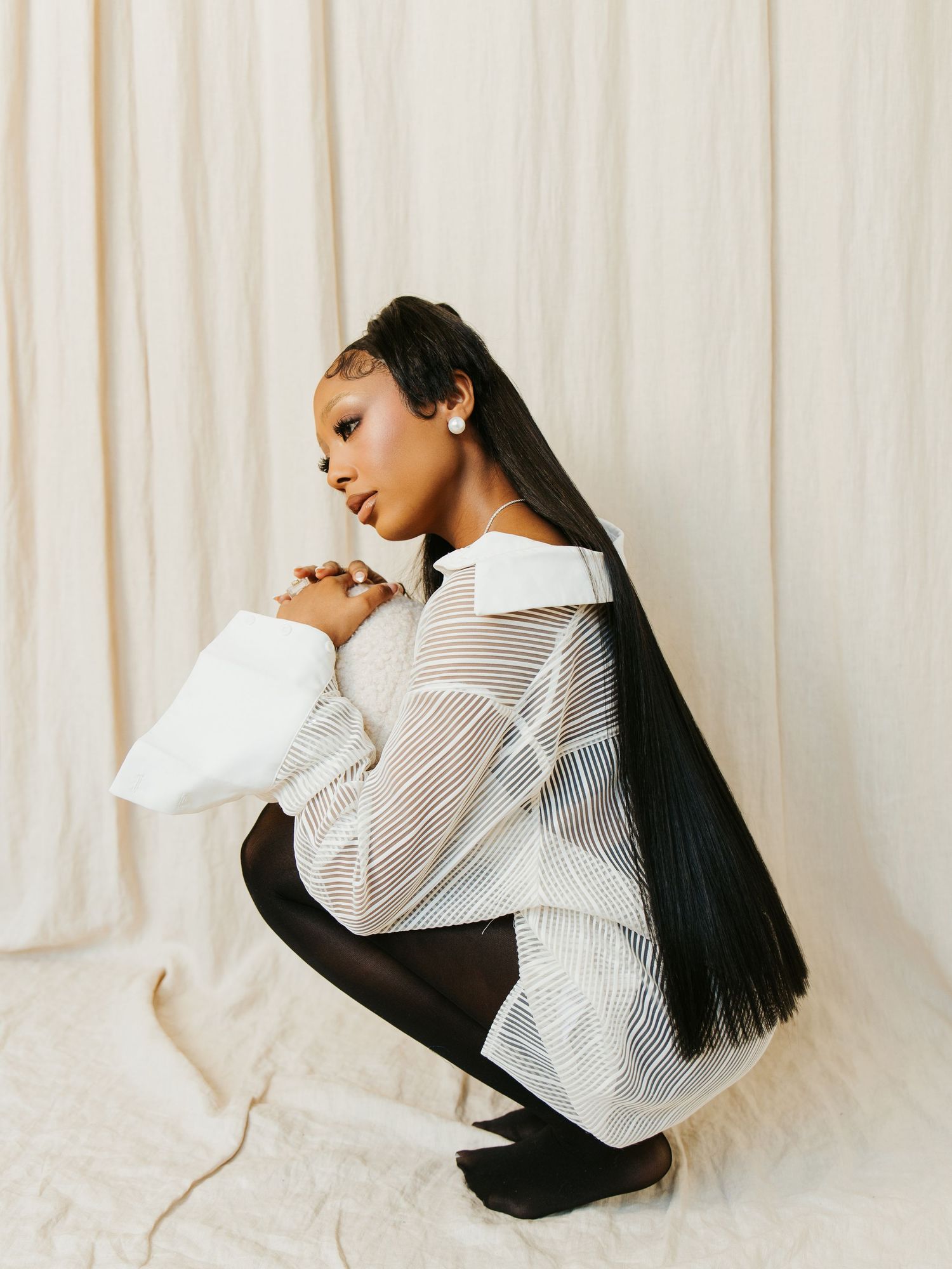 A woman in a sheer white top and black tights crouches gracefully against a neutral fabric backdrop.