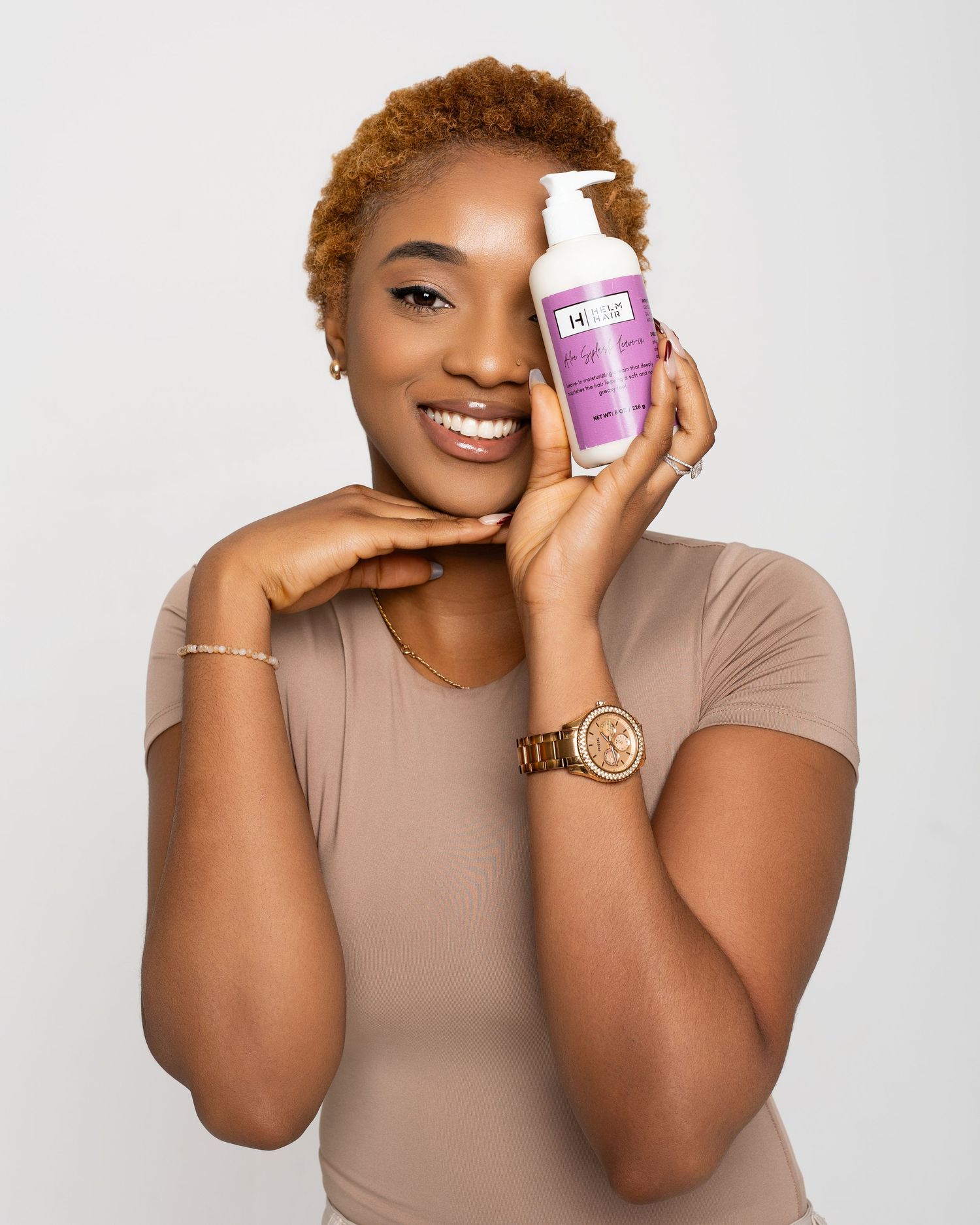 Smiling woman holding a purple and white skincare product bottle against her face, wearing a beige top and gold watch.