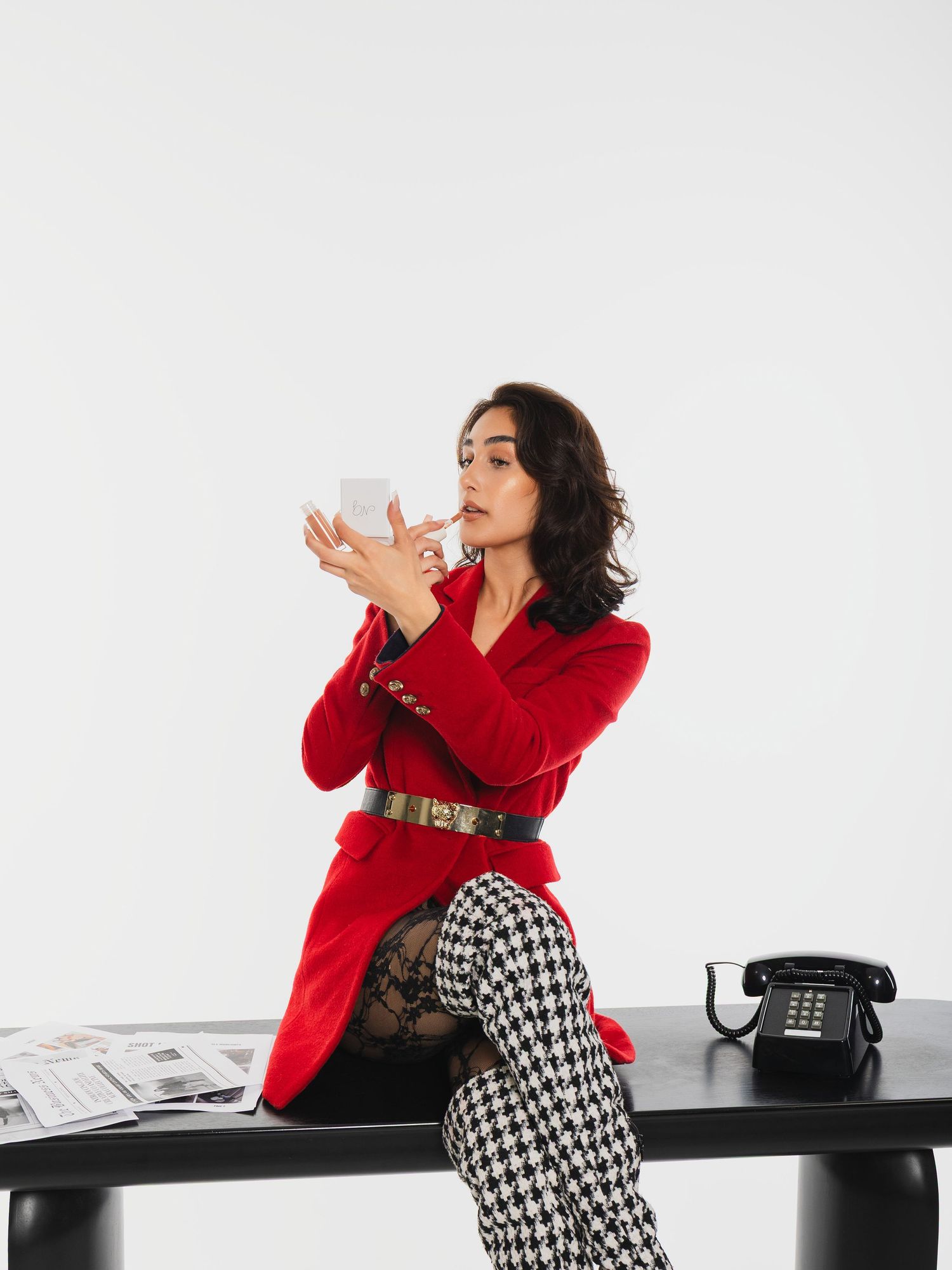 Woman in red blazer and houndstooth pants sits on desk, holding white cup near vintage phone and newspaper.