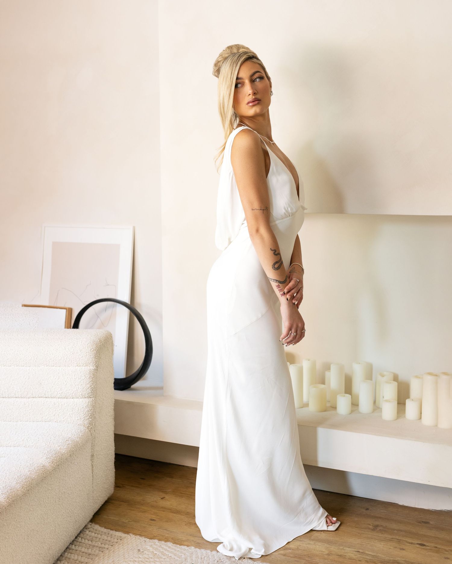 Woman in elegant white sleeveless maxi gown posing sideways in a modern minimalist room with candles and neutral decor.