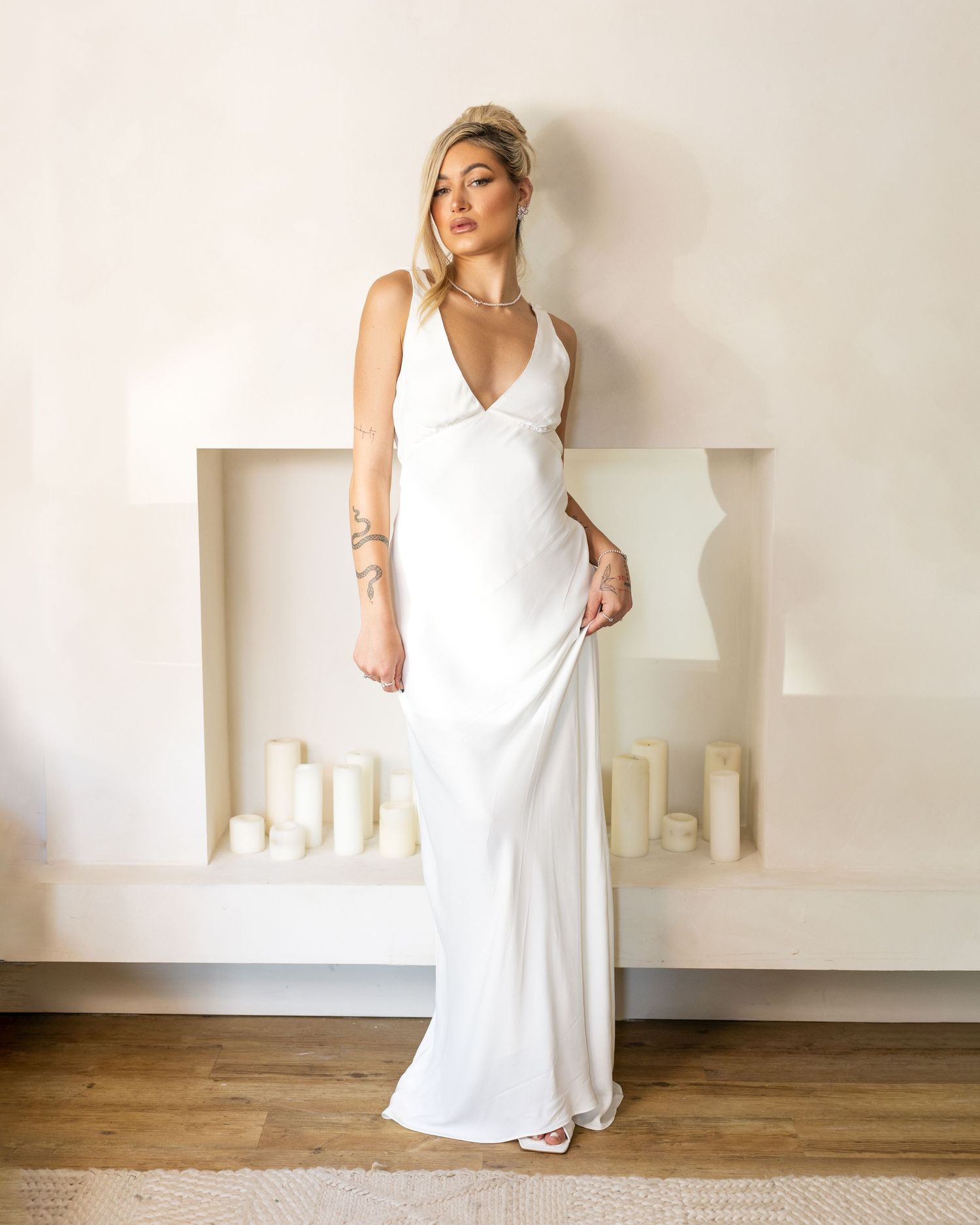 Woman wearing a white satin maxi slip dress with V-neckline, posing in front of a minimalist white fireplace.