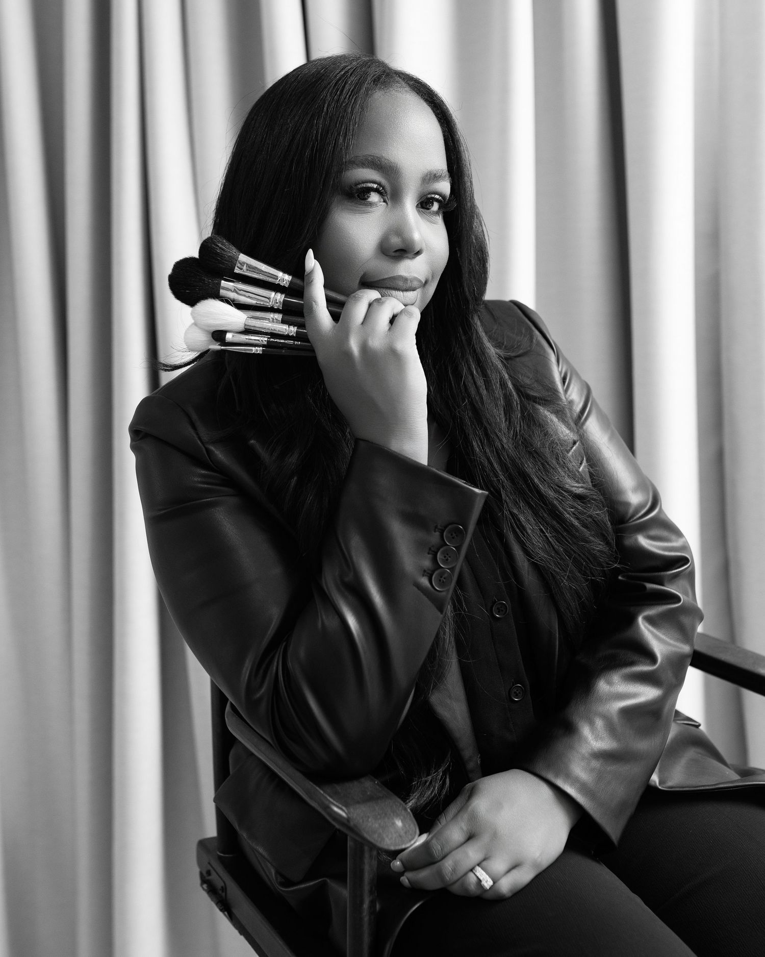 Black and white portrait of a woman in leather jacket holding makeup brushes, posing thoughtfully against draped curtain backdrop.