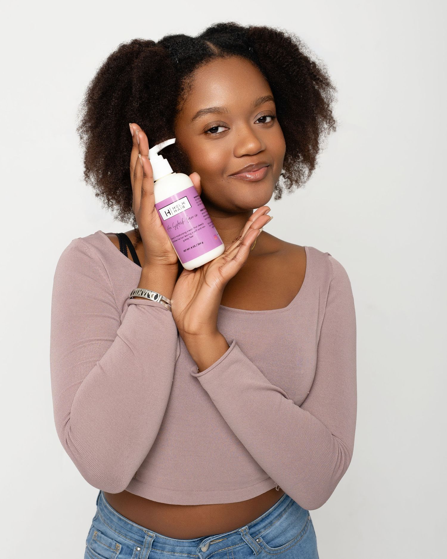 Young woman with natural hair holding a purple pump bottle skincare product against a light background.