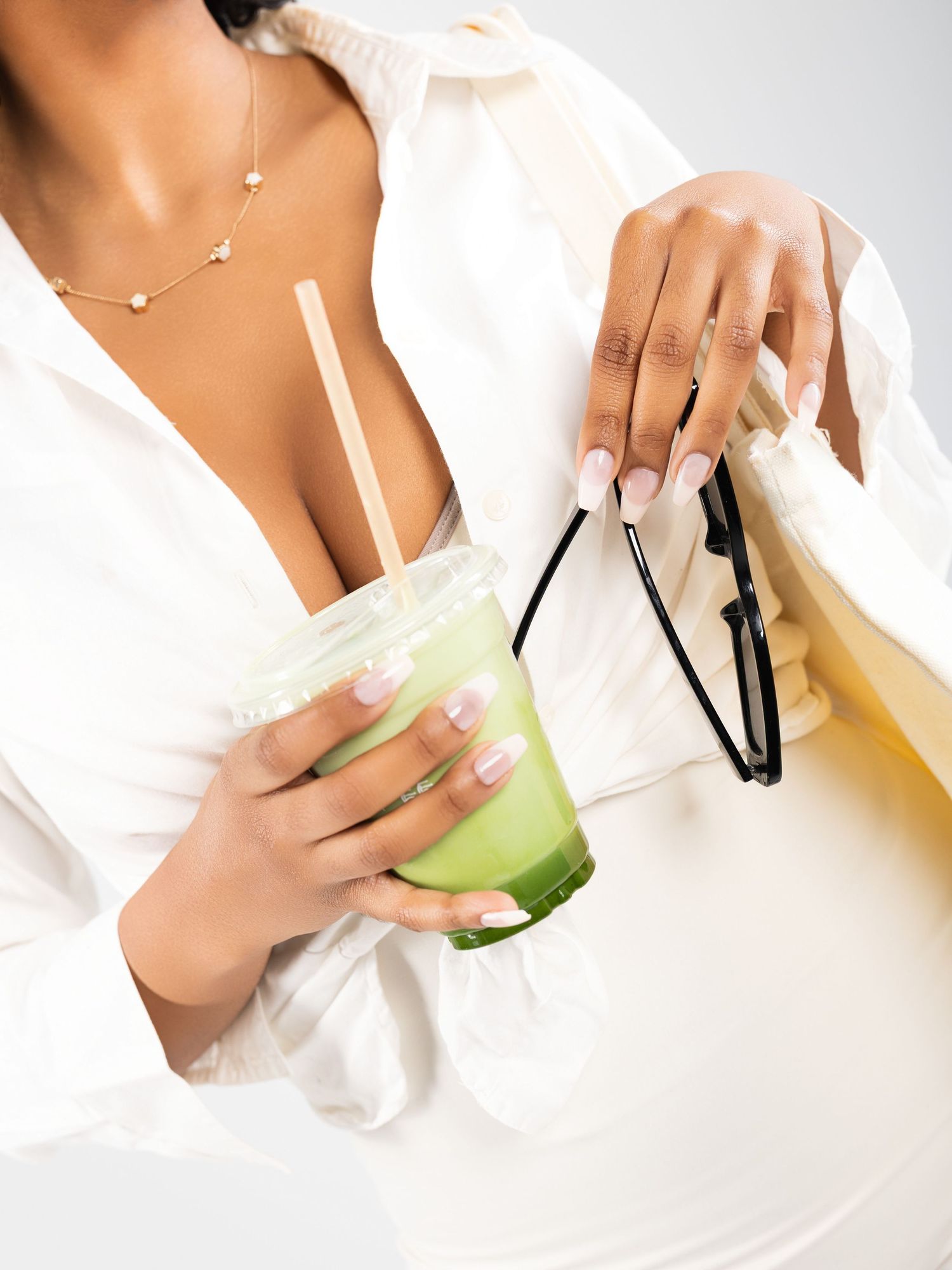 Woman in white shirt holding iced matcha latte and sunglasses, wearing gold necklace, with manicured nails.