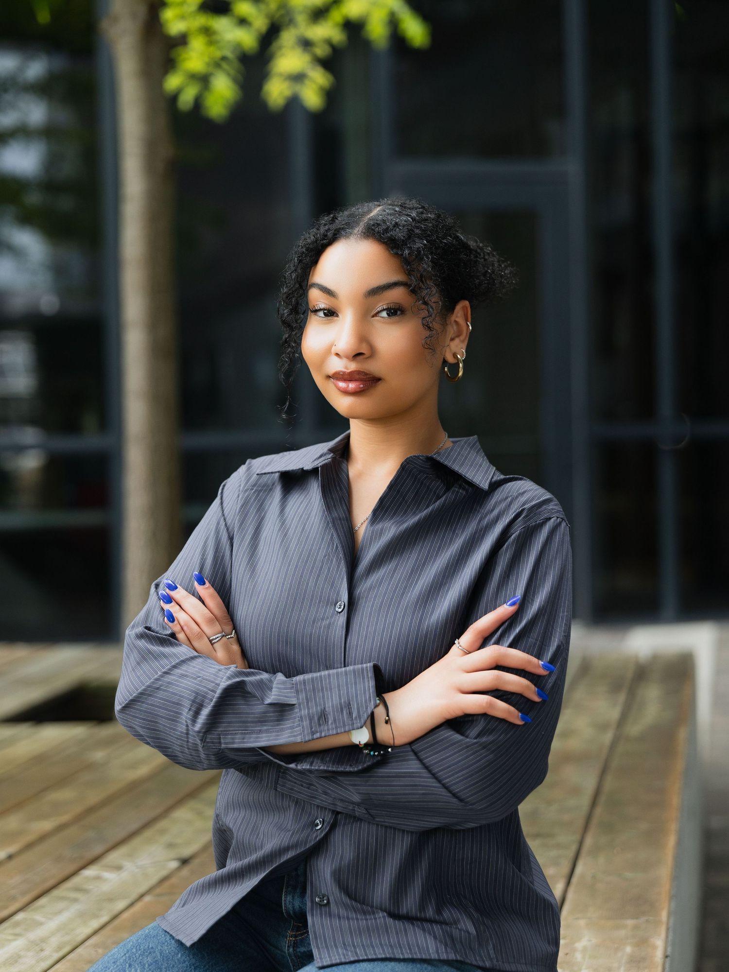 Young woman in gray striped shirt sits with arms crossed outdoors, exuding confidence near modern building.