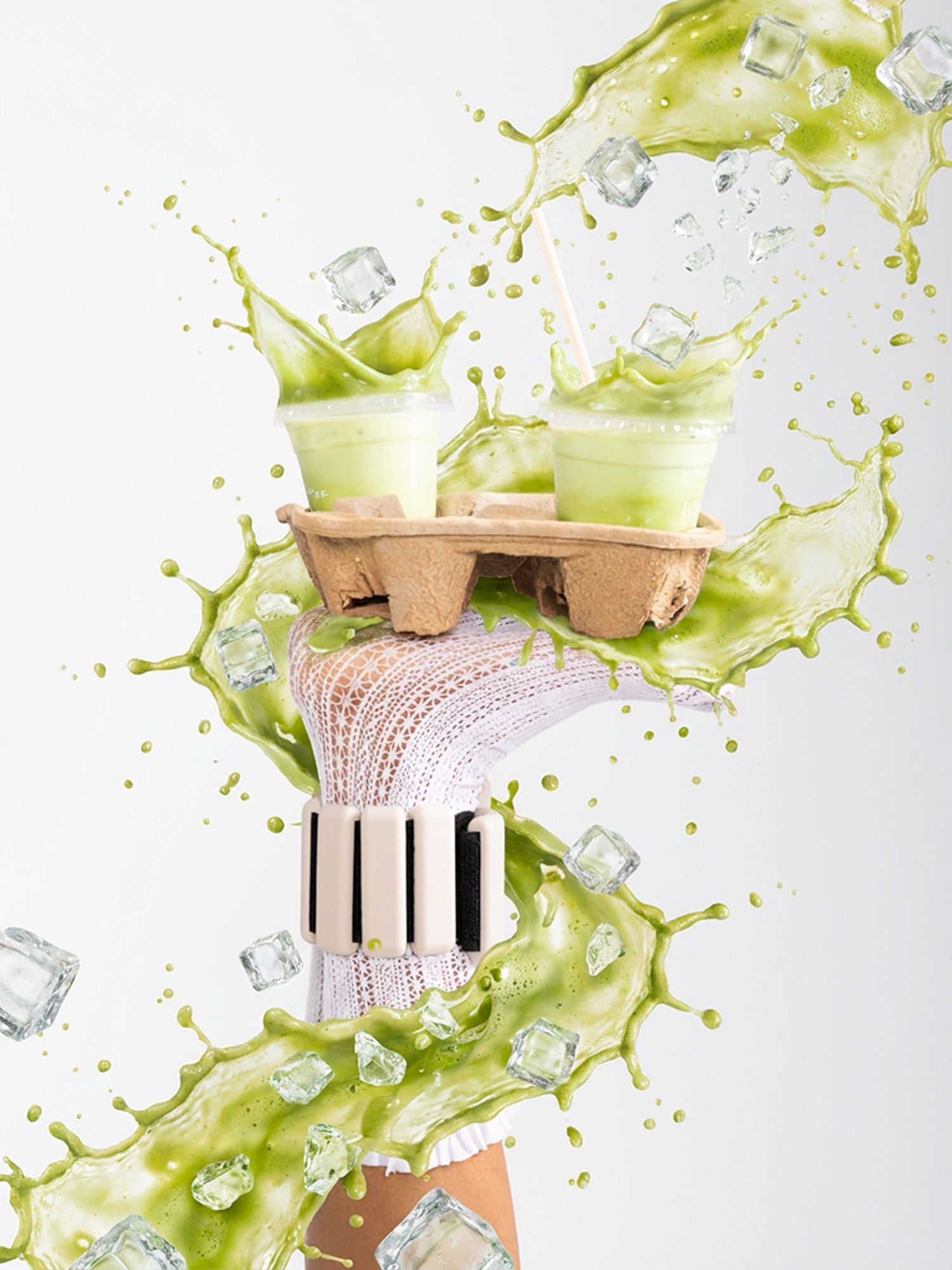 Hand holding two matcha green tea drinks in a cardboard carrier with ice cubes and green liquid splashing dramatically.