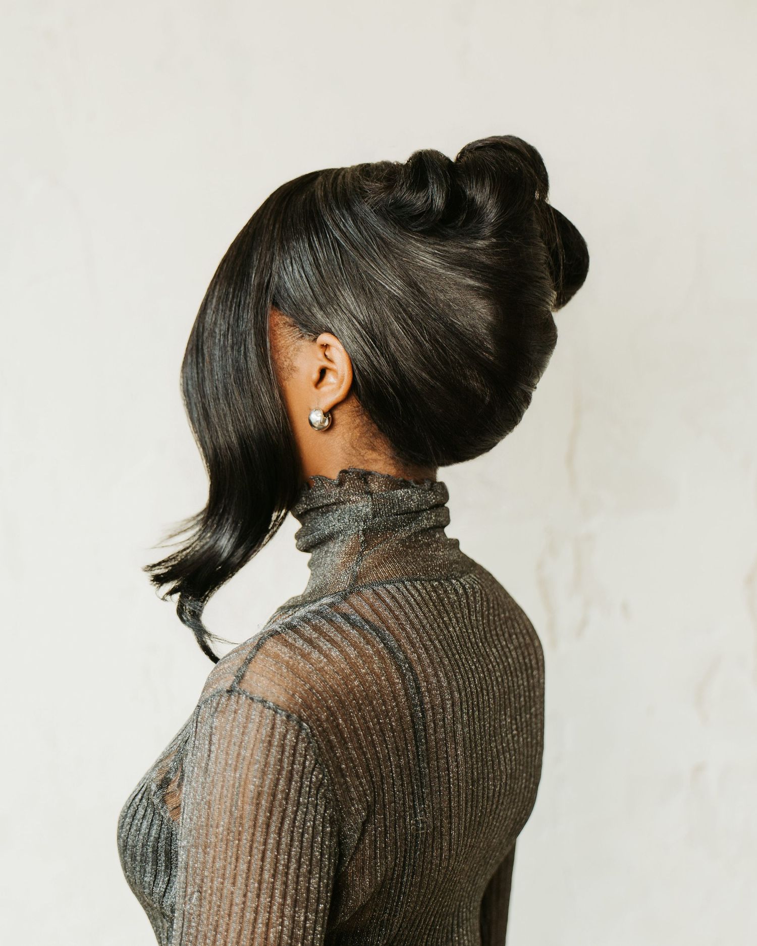 Side profile of a Black woman with elegant updo hairstyle wearing a sheer turtleneck top against white wall.