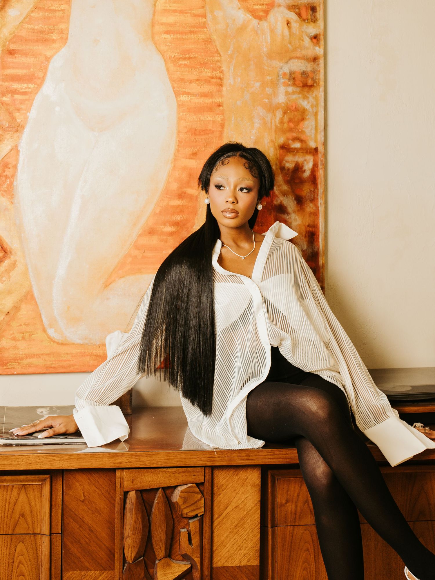 A woman in a white sheer blouse, black shorts and tights sits on a wooden dresser with long straight hair.