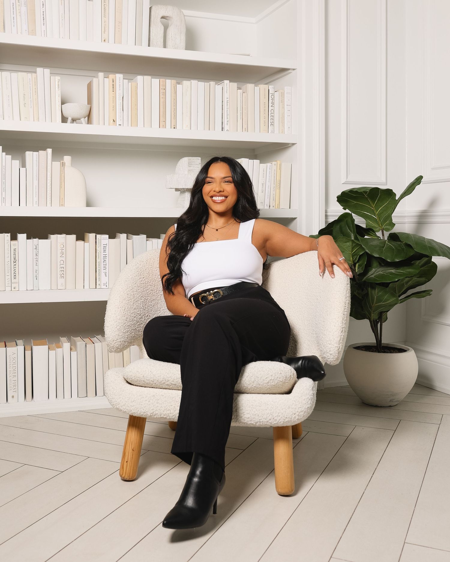 Woman in white top and black pants sitting in a cozy chair, surrounded by white bookshelves and a green plant.