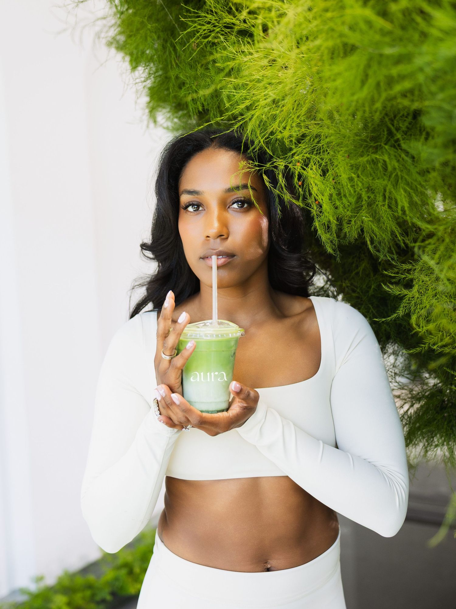 Woman in white crop top holding a green matcha smoothie drink with a straw against lush green foliage backdrop.