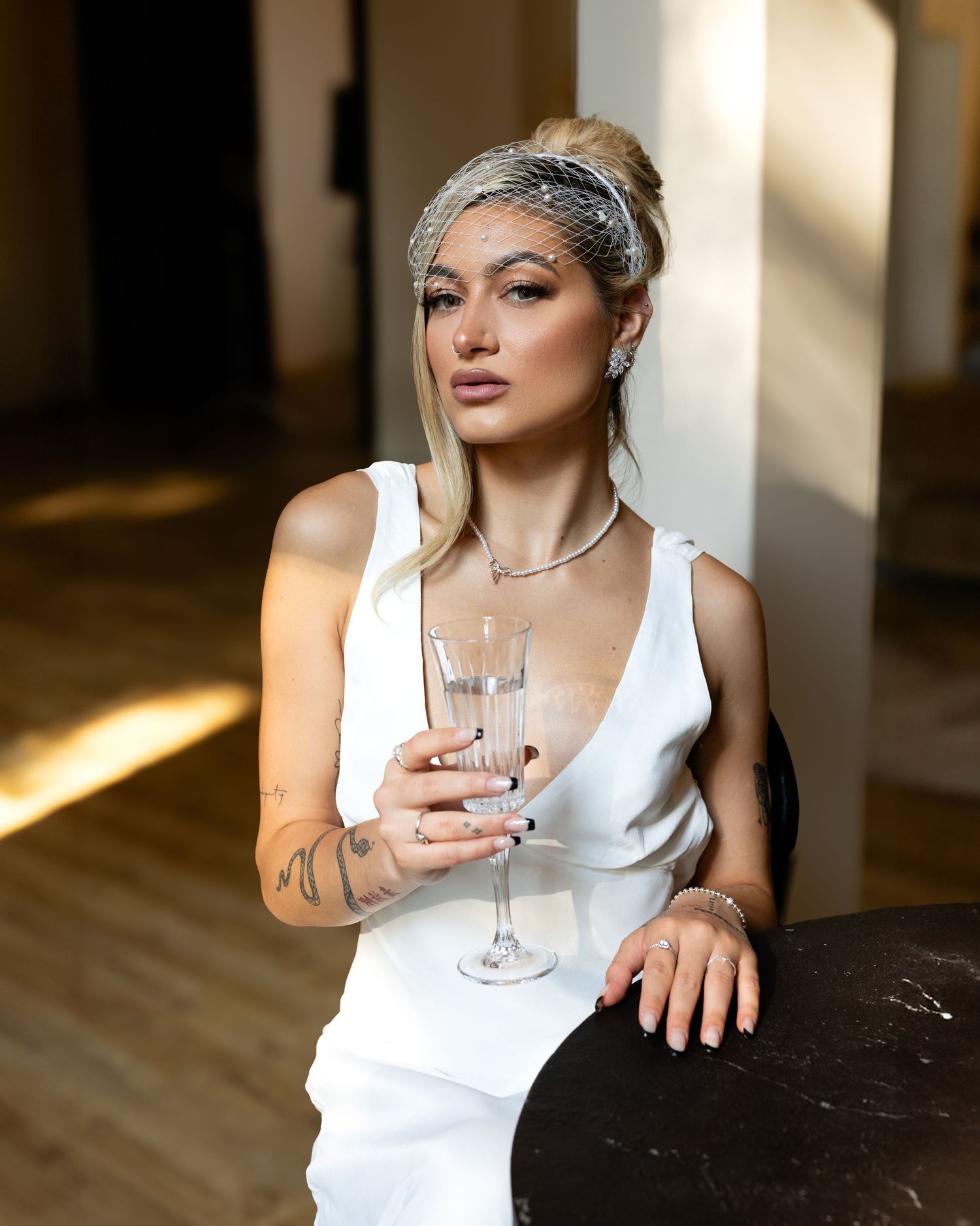 A woman in a white dress holds a champagne flute, wearing a veil headband and tattoos, posed elegantly indoors.