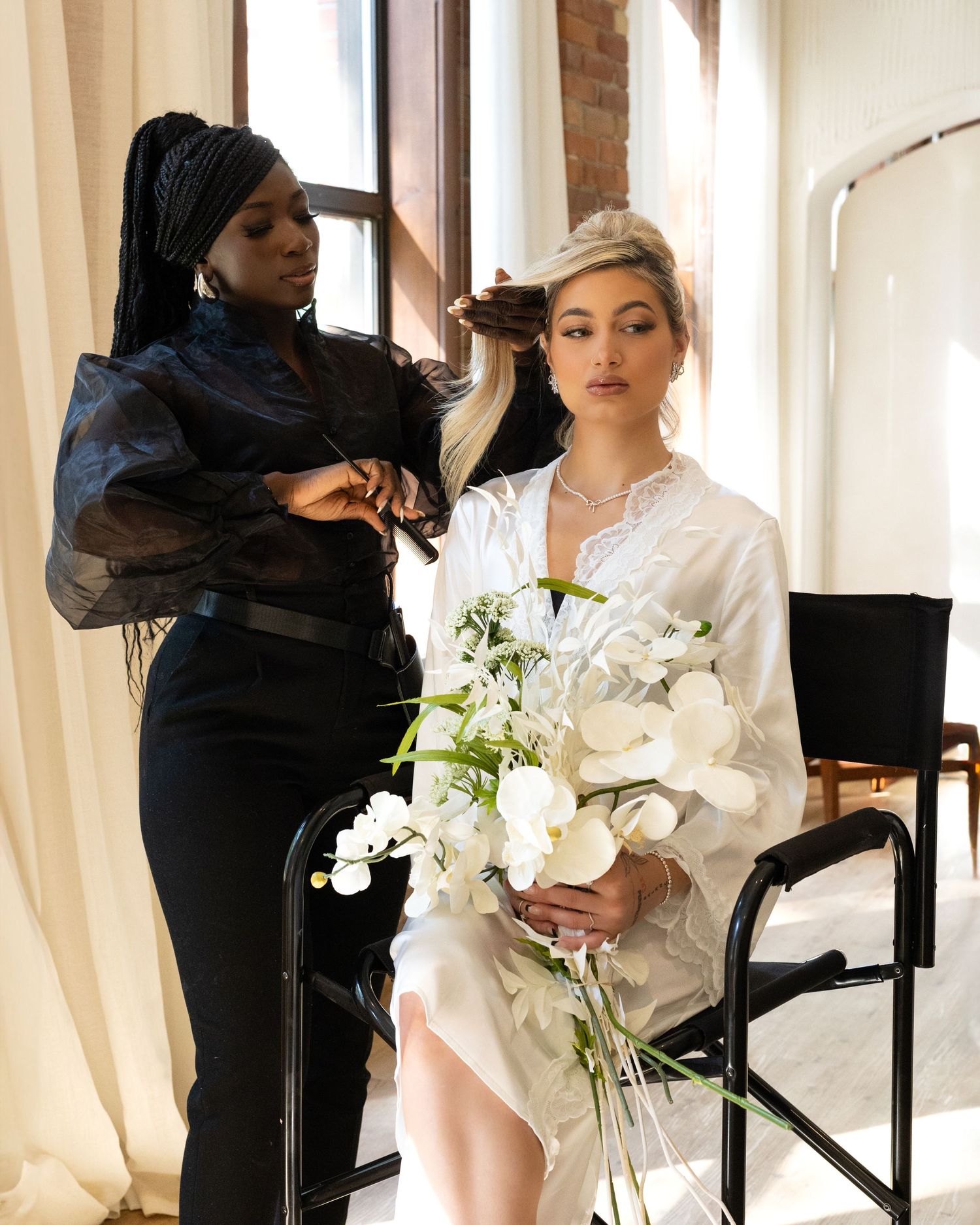 Bride in white robe holds bouquet of white flowers while stylist does her hair in bright studio.