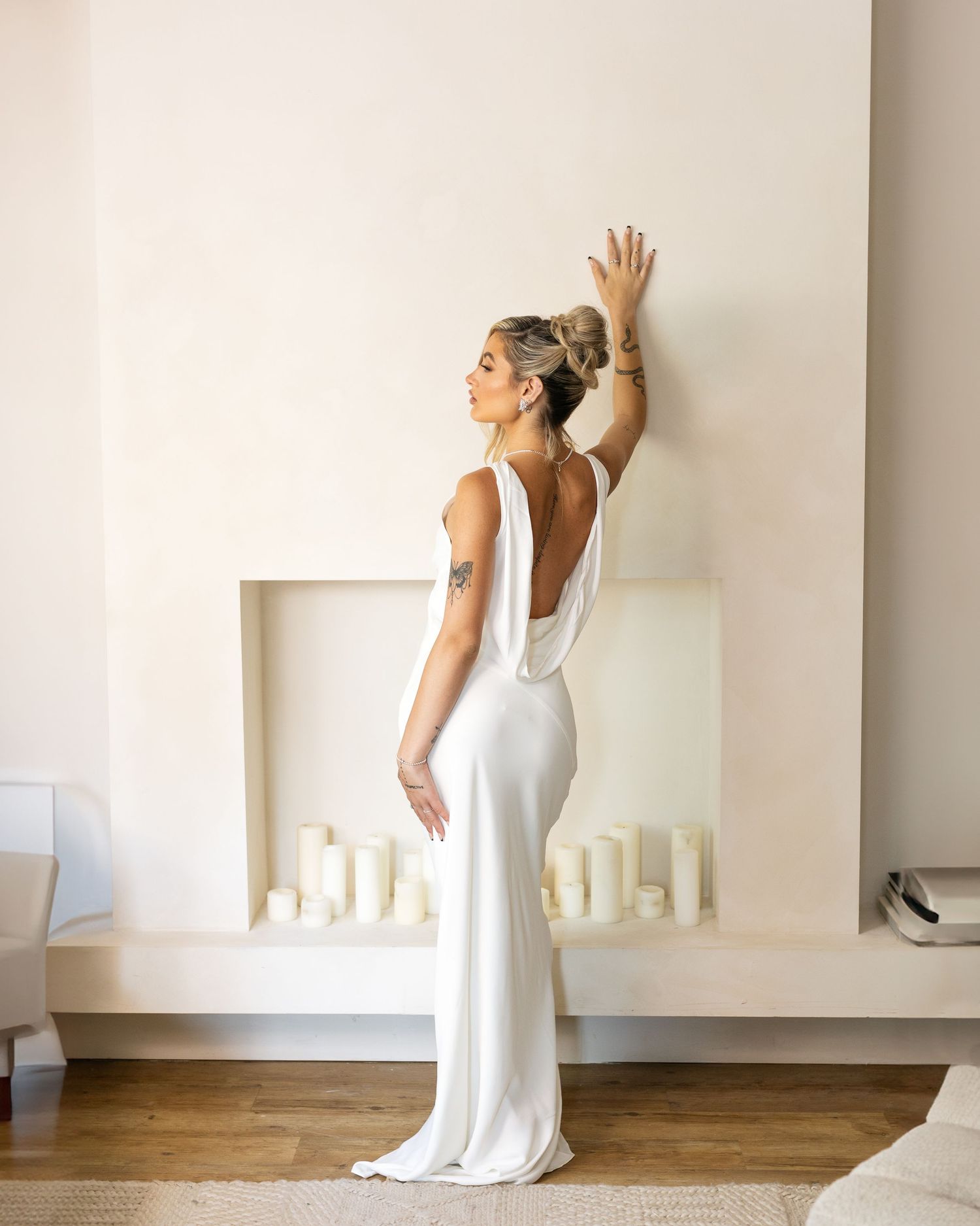 Woman in elegant white backless satin gown poses gracefully against minimalist white wall with candle fireplace.