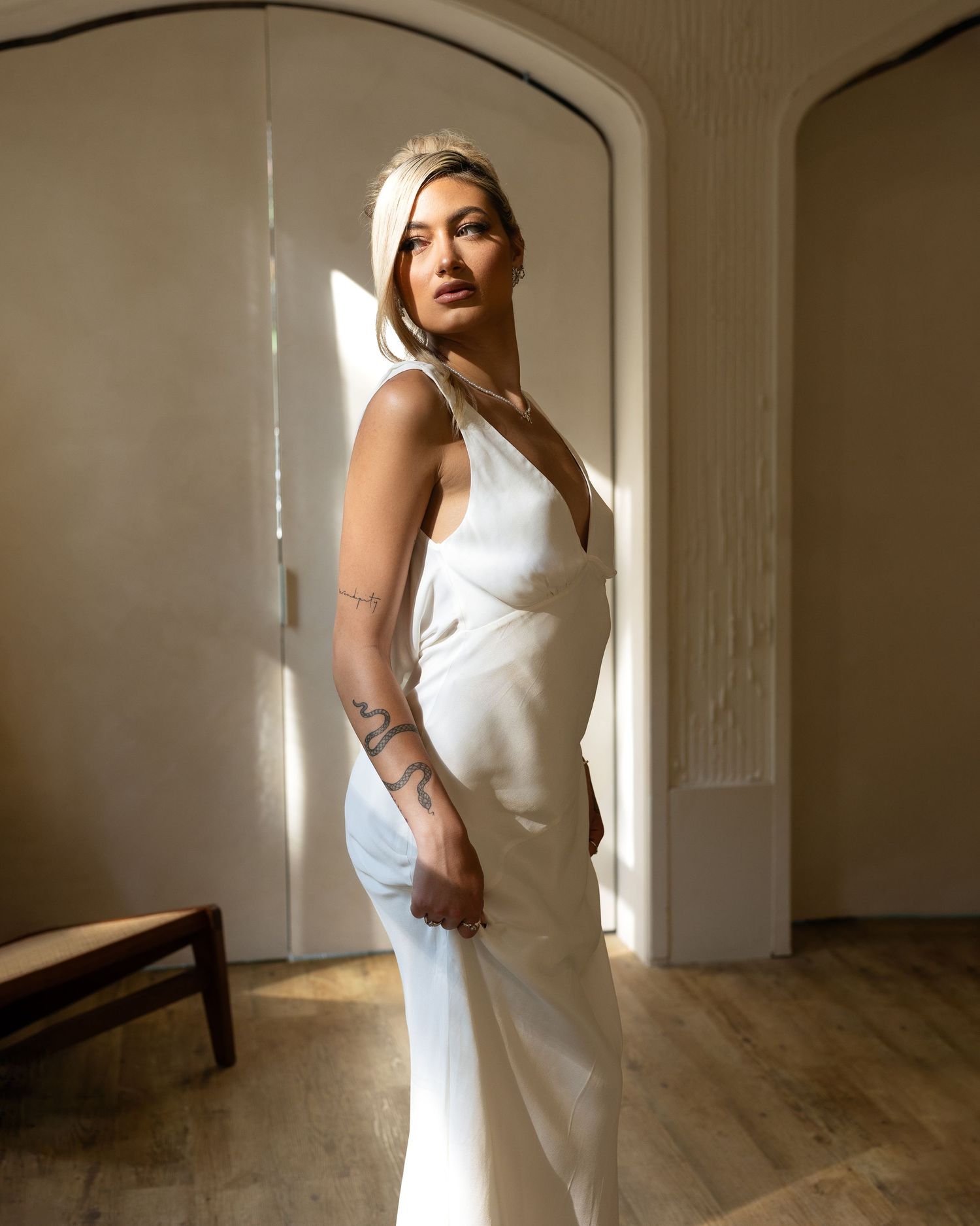 Woman in white satin slip dress with tattoos poses in sunlit room with arched doors and hardwood floors.