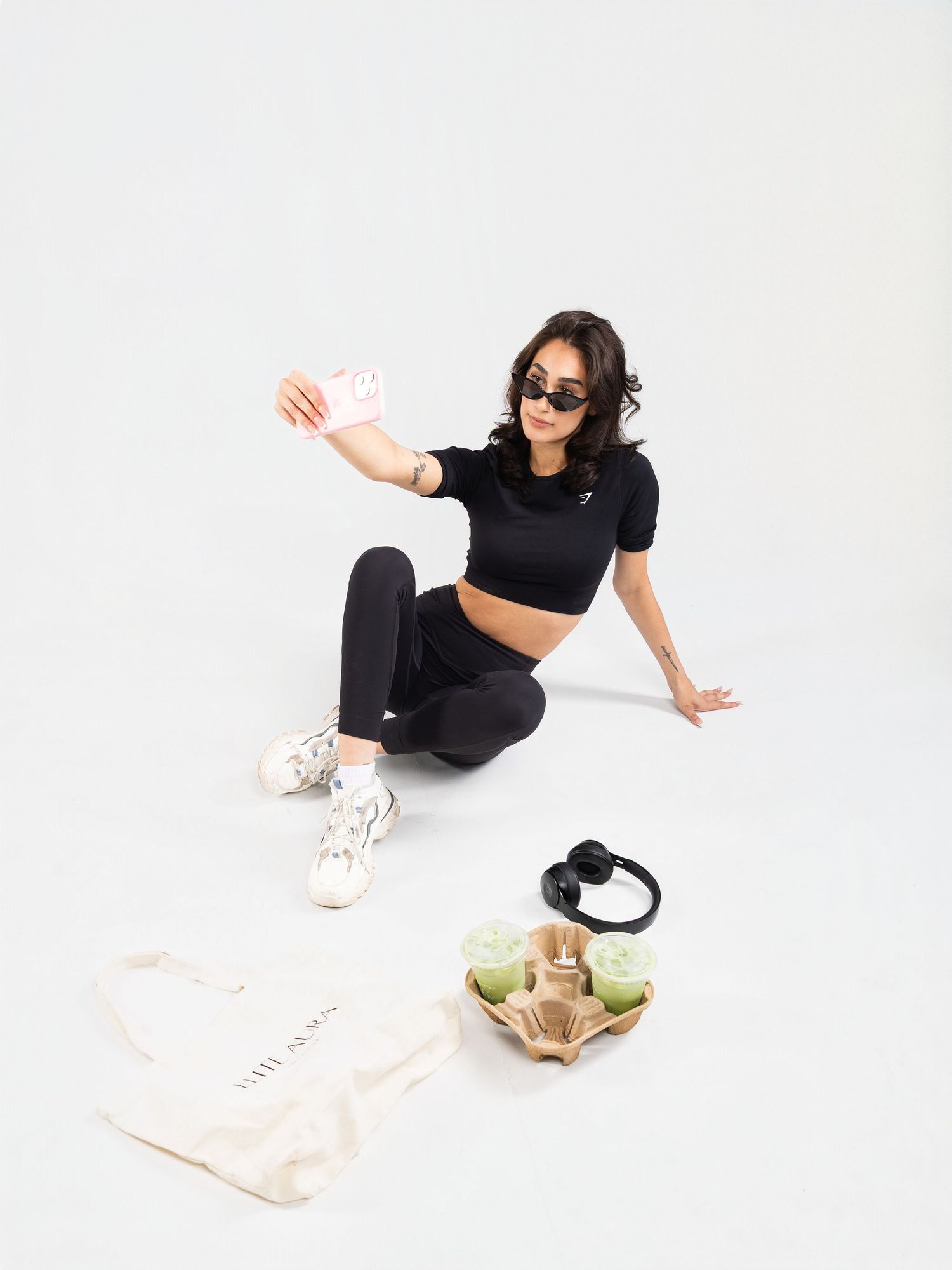 Woman in black crop top and leggings taking a selfie, seated with tote bag, headphones, and green drinks nearby.