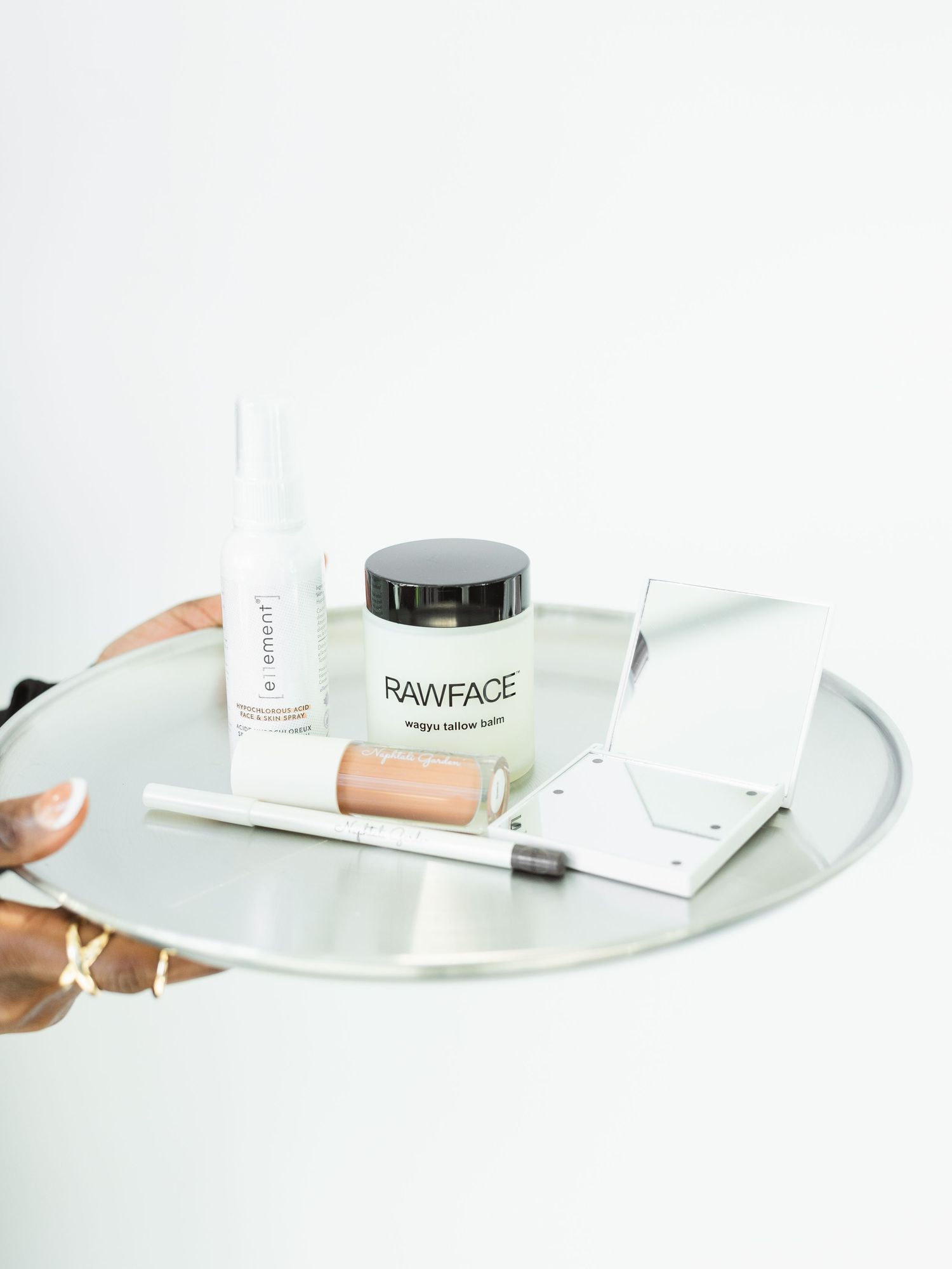 A silver tray holding Rawface tallow balm, a lip gloss, serum, and makeup brush displayed on a white background.