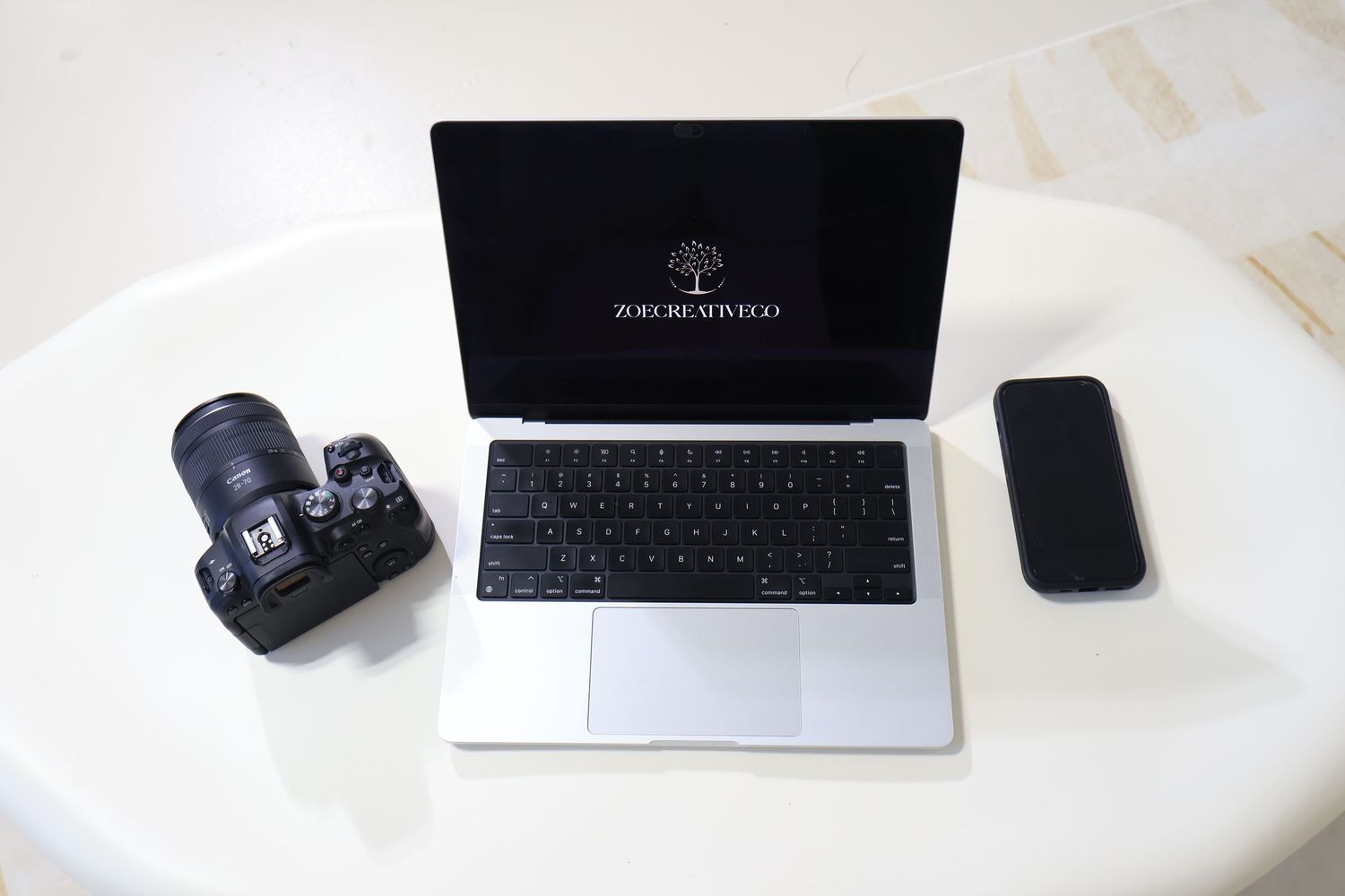 A MacBook laptop, DSLR camera, and smartphone arranged on a white round table in a flat lay composition.