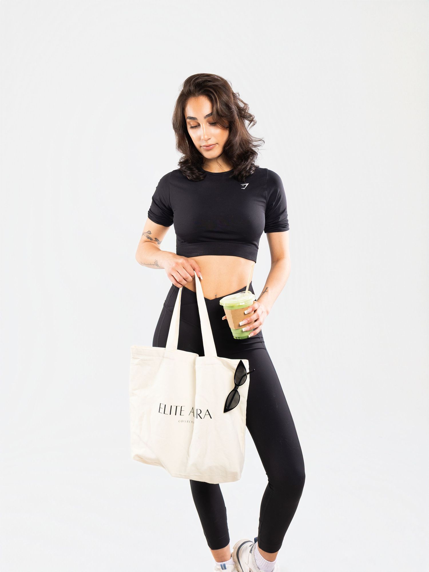 Woman in black crop top and leggings holding a green smoothie and Elite Aura tote bag.