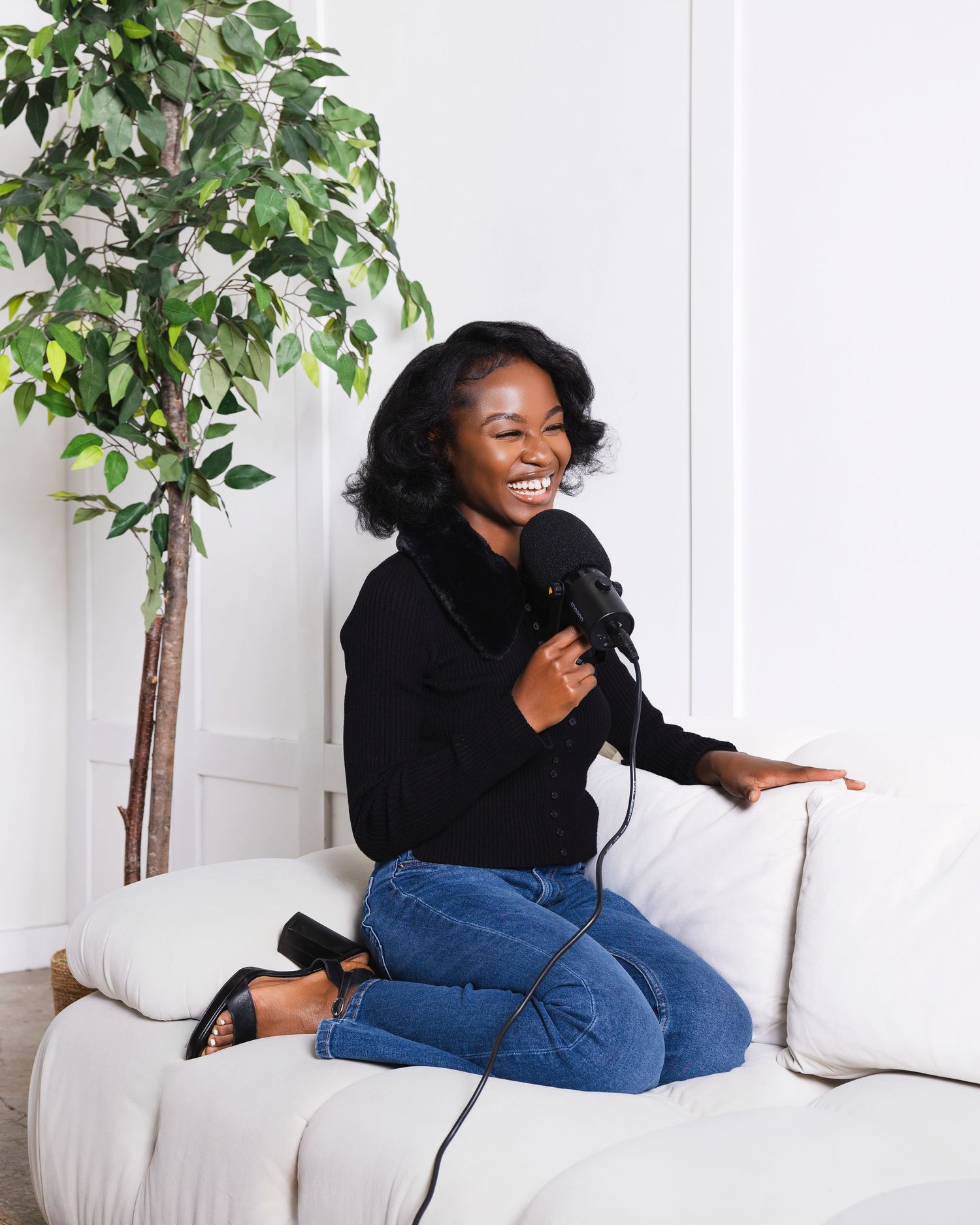A smiling Black woman sits cross-legged on a white sofa holding a podcast microphone, wearing jeans and a black top.