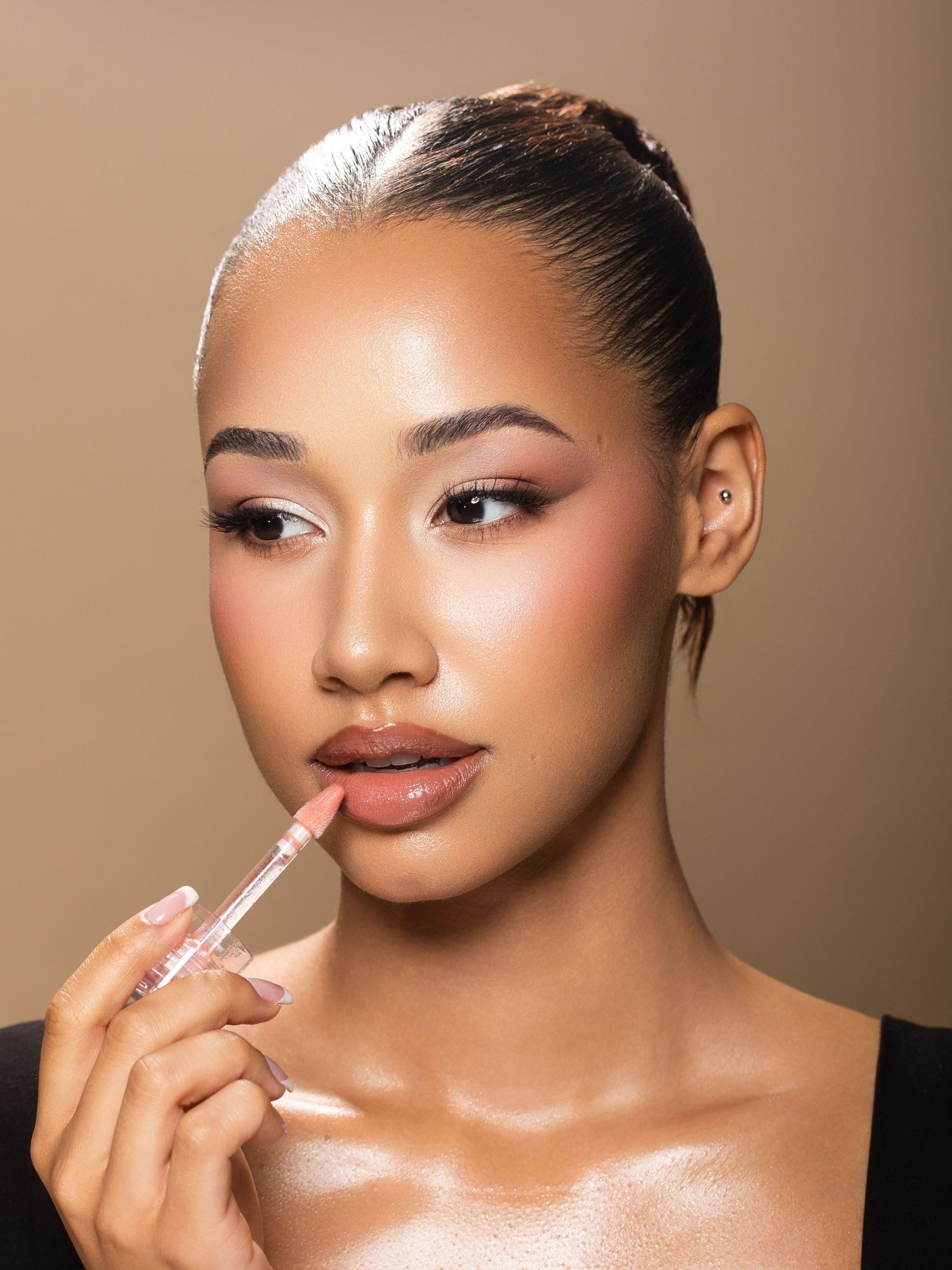 A young woman applies clear lip gloss with an applicator wand, showcasing a glowy makeup look.