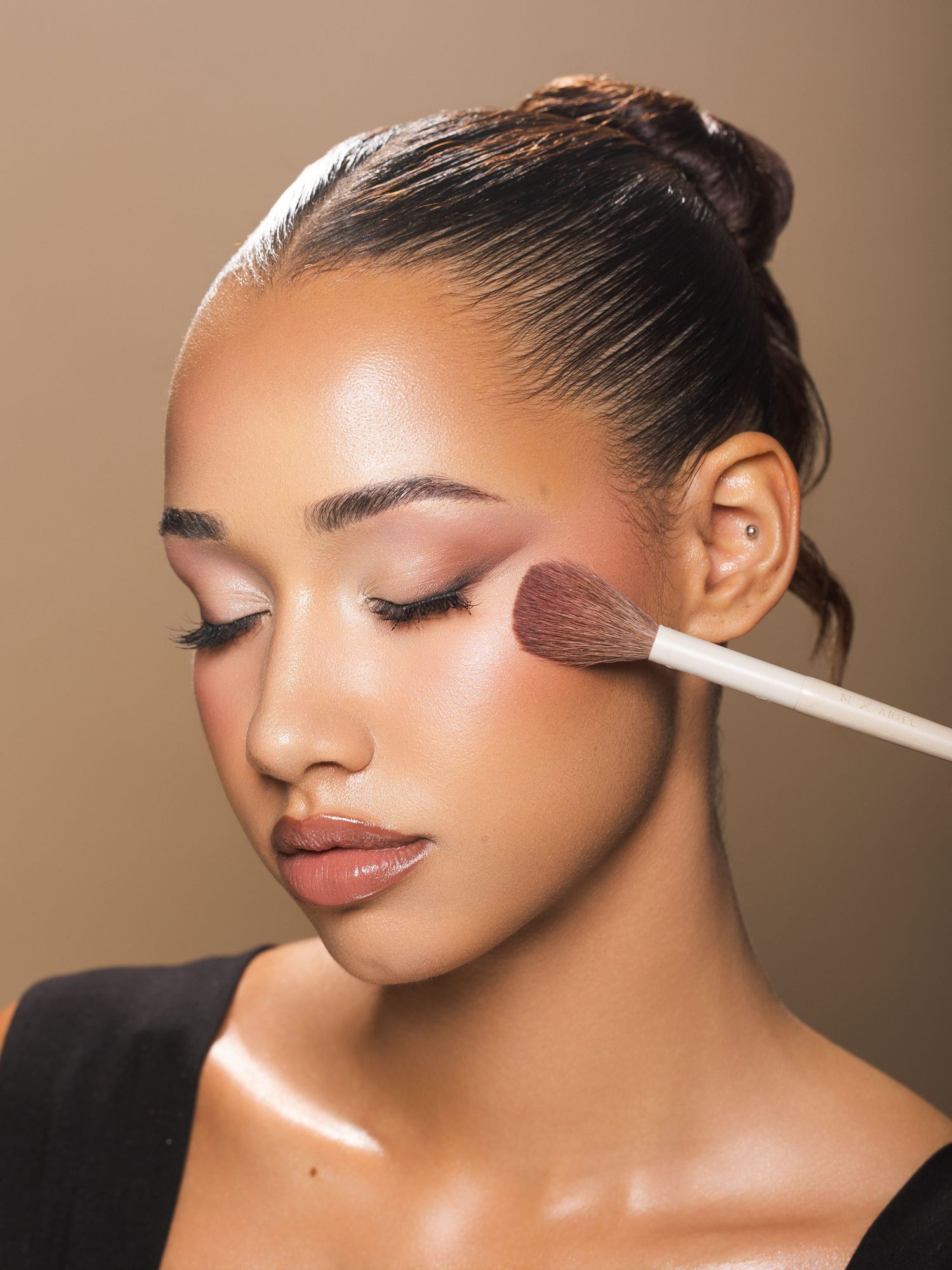 A makeup artist applies blush with a white brush to a young woman with slicked-back hair and natural glam makeup.
