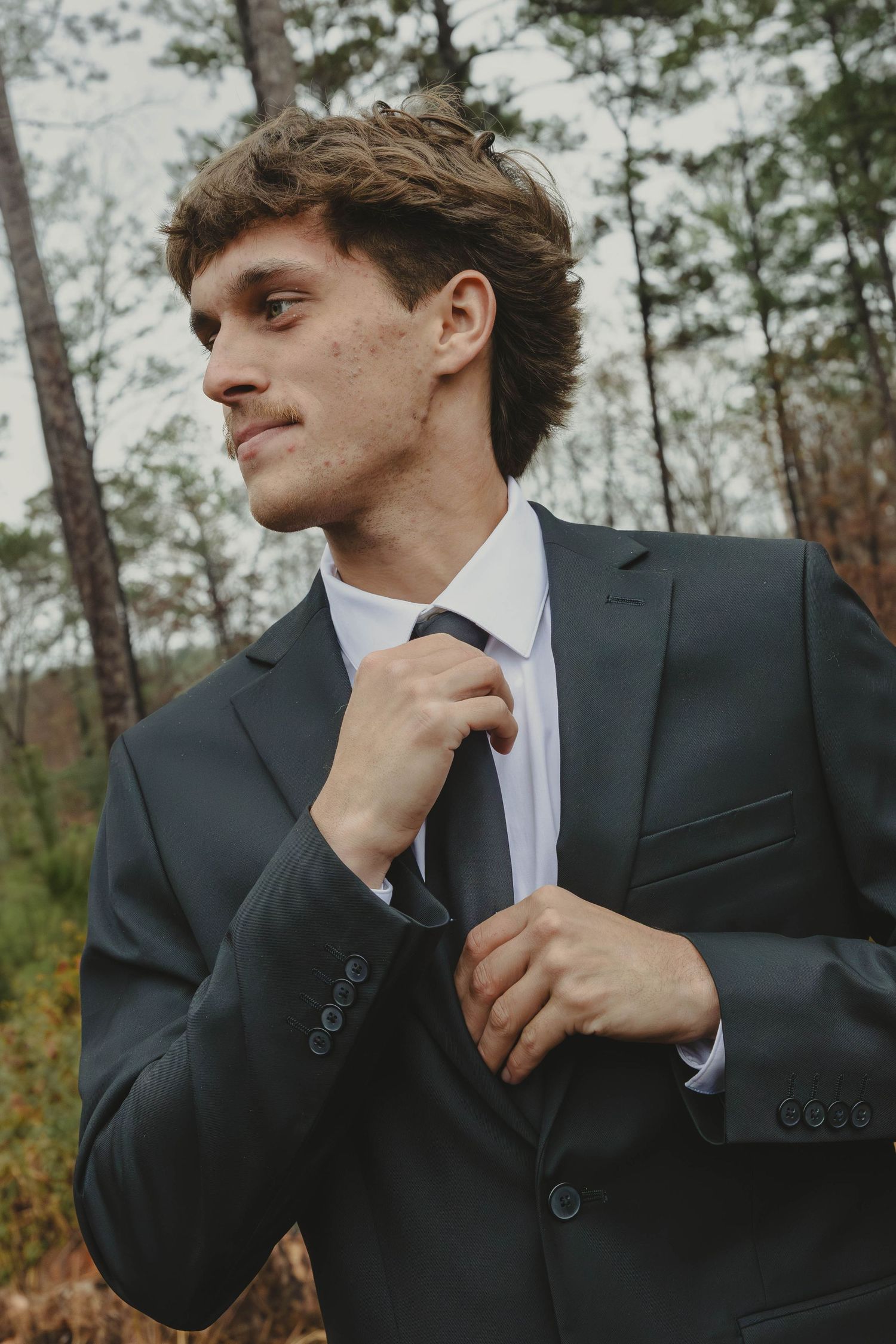 A person in a dark suit and white shirt poses thoughtfully against a pine forest background.