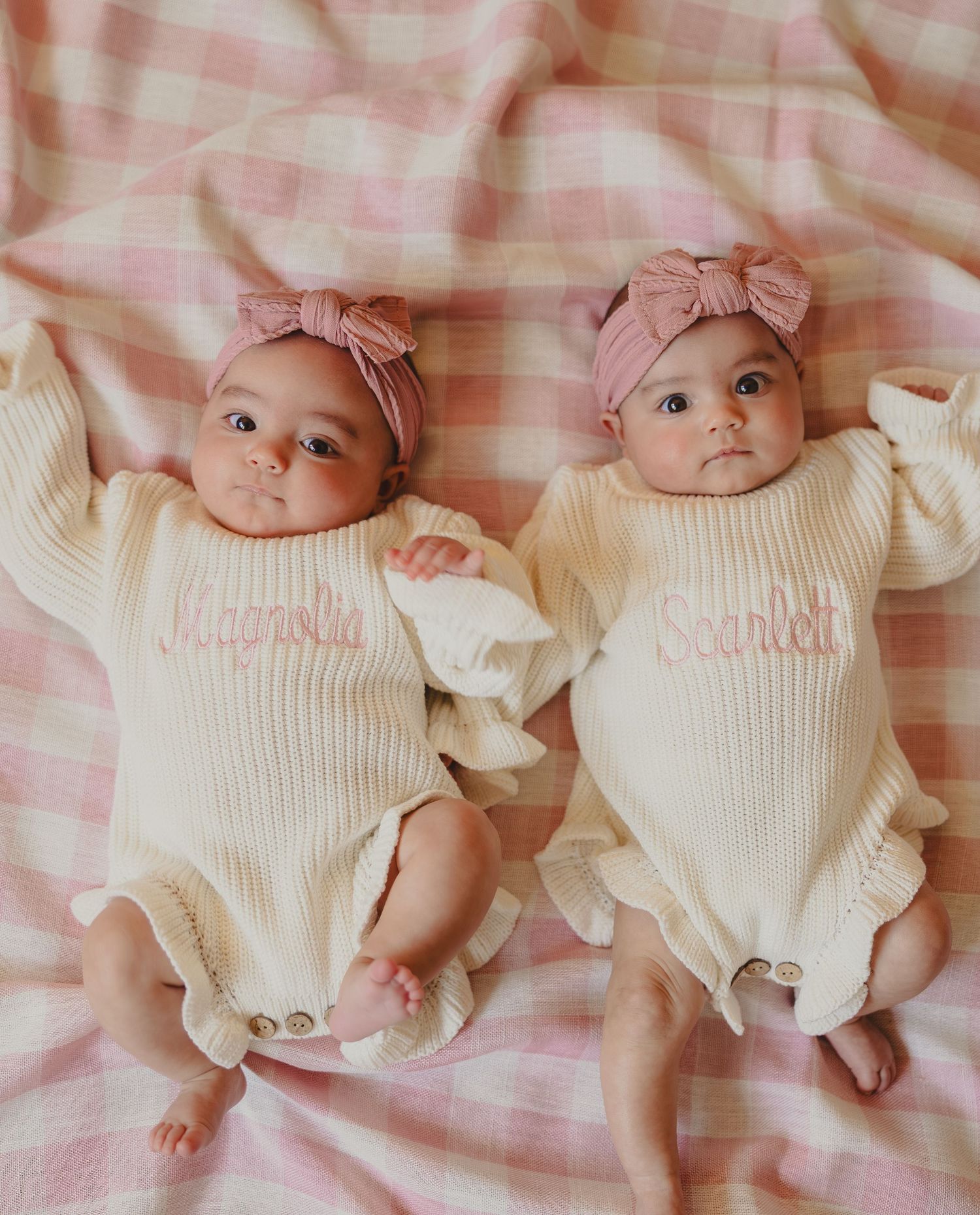 Newborn twins in matching cream-colored outfits lying on pink and white checkered bedding.