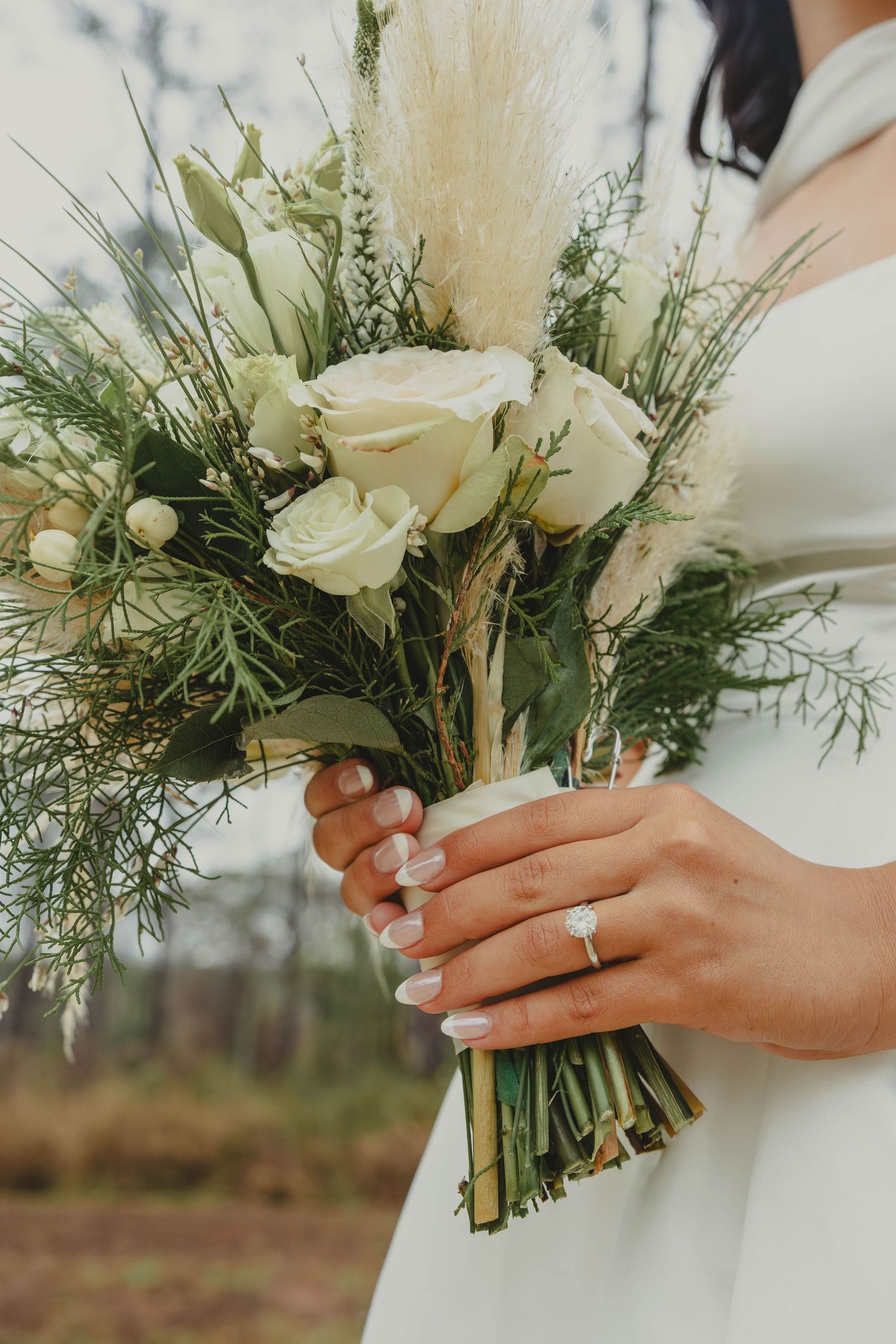 Close-up of hands holding an elegant white rose and greenery wedding bouquet with a sparkling engagement ring.