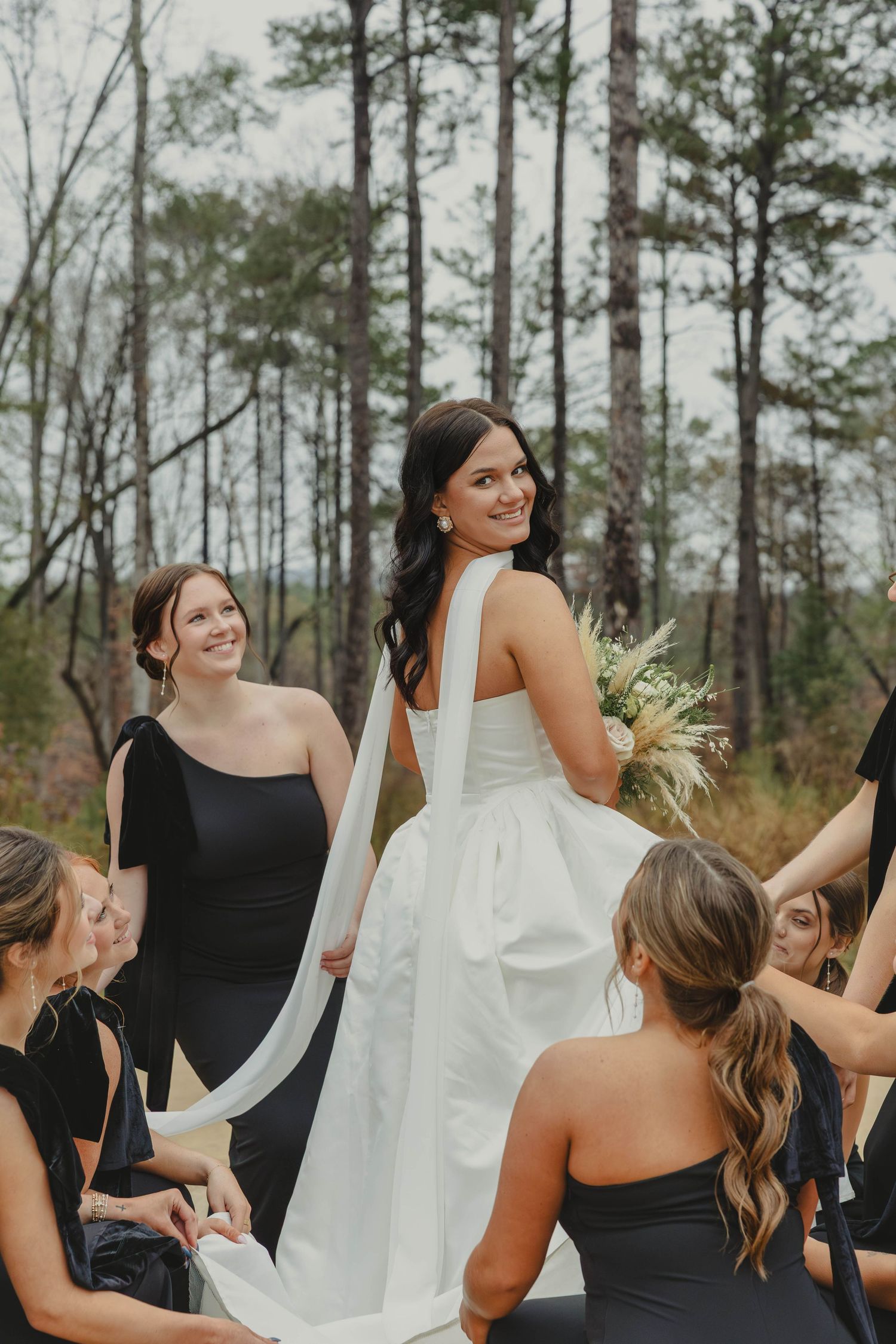 Wedding party wearing black dresses stands together outdoors among tall pine trees.