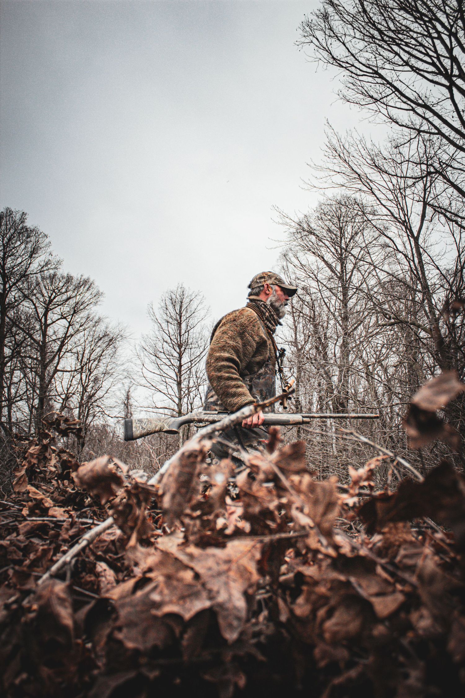 Hunter in camouflage gear walking through autumn forest with fallen leaves and bare trees.