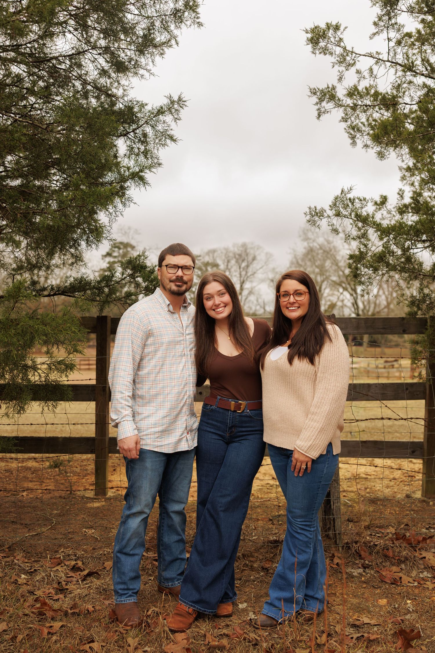 Three people stand together outdoors on fallen leaves against a wooden fence and trees in autumn.