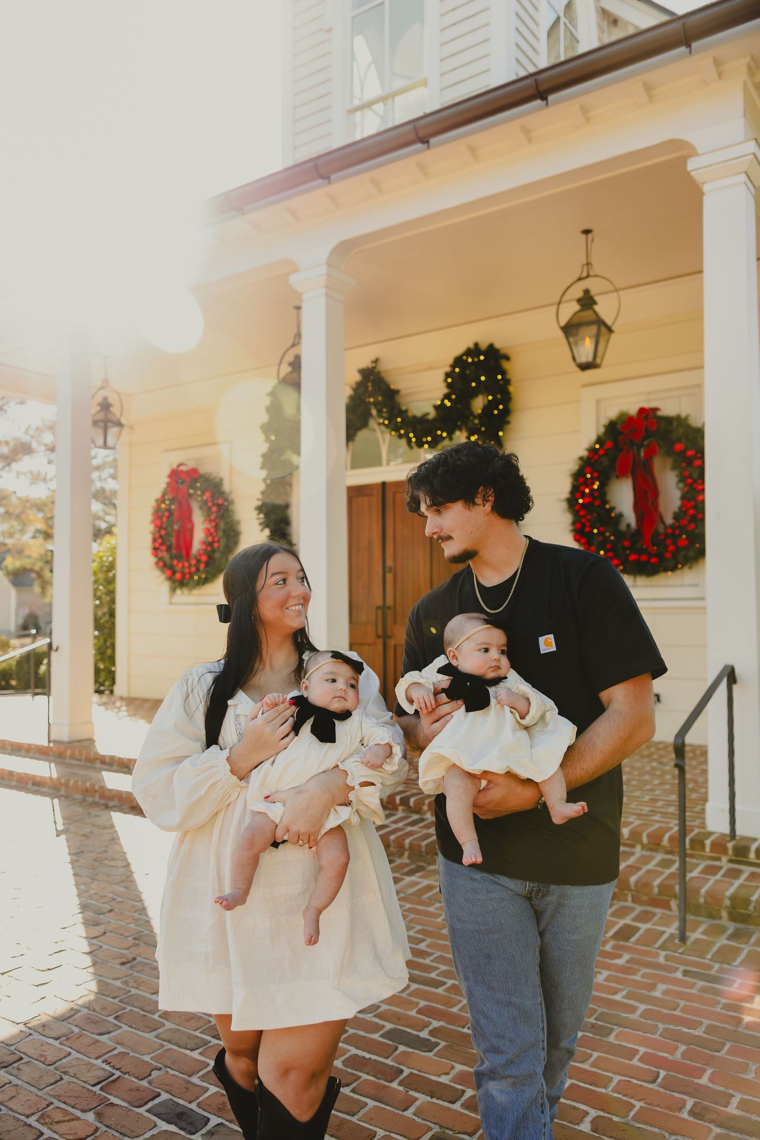 A couple stands on a brick porch decorated with Christmas wreaths while holding twin babies in their arms.